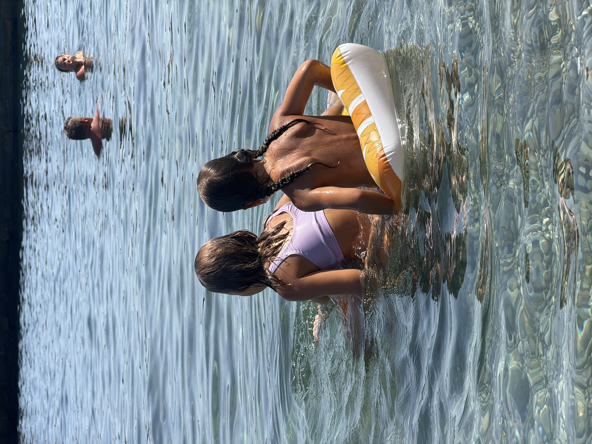 The image depicts a group of people enjoying a sunny day in clear, shallow water. Two young girls are the primary subjects, positioned in the foreground. The girl on the left is wearing a purple swimsuit and appears to be reaching out. The girl on the right, wearing a bikini top and bottom, is leaning back against an inflatable ring. Her hair is styled in two braids. In the background, two individuals are partially submerged in the water, with their heads and shoulders visible. The water is calm and transparent, allowing a view of the pebbles on the seabed. Sunlight reflects off the water's surface, creating a shimmering effect. The scene suggests a leisurely activity, likely swimming or playing in the water on a warm day. The environment appears to be a beach or a swimming area. The bright lighting indicates it is daytime. There is no visible text in the image.
