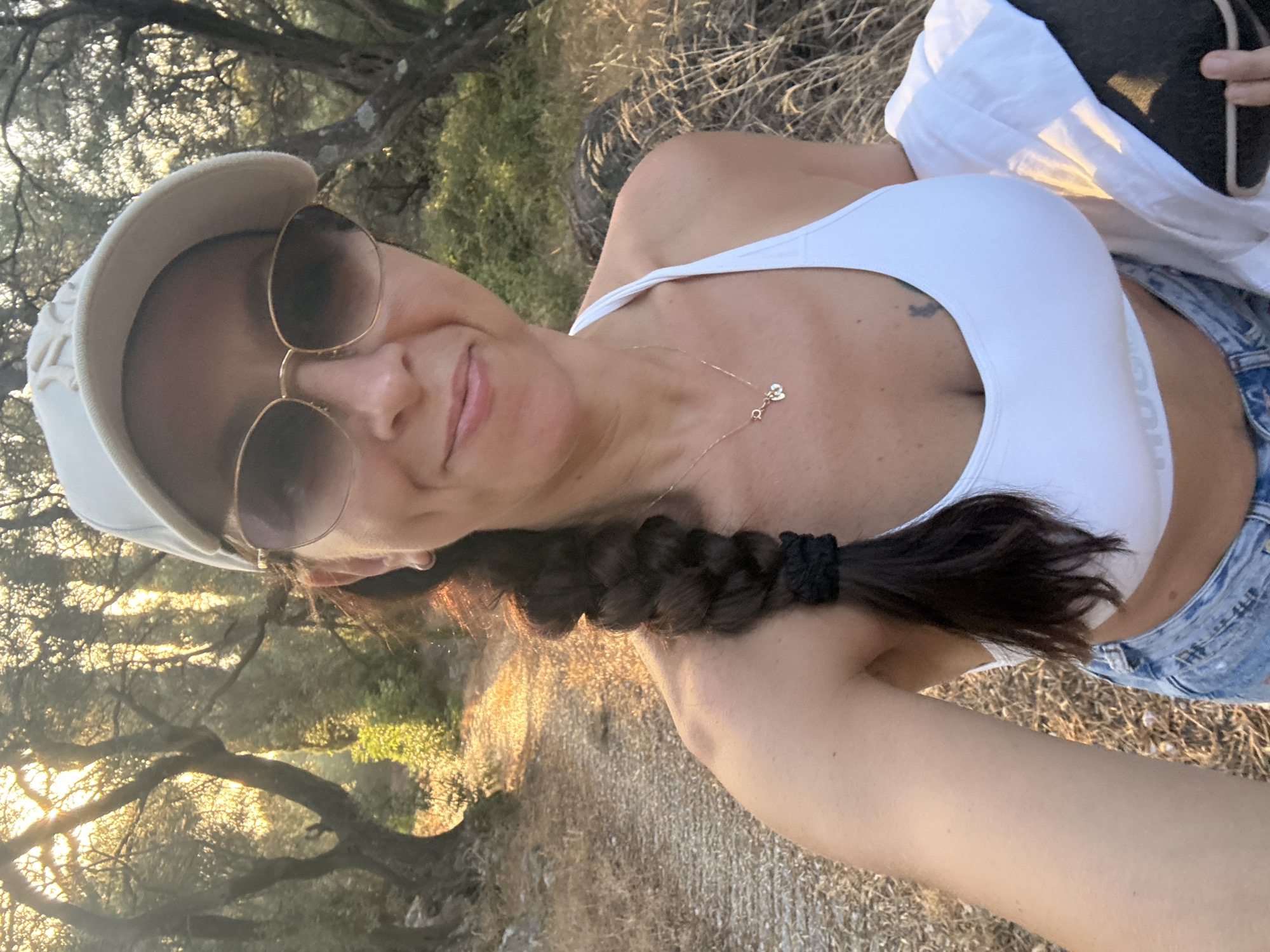 A woman is taking a selfie in a sun-dappled wooded area. She is wearing a white sports bra, denim shorts, sunglasses, and a baseball cap. Her hair is in a braid. She is holding a white shirt and a dark object in her right arm. The background shows a path lined with trees and dry grass. The scene appears to be outdoors in a rural or natural setting, possibly Makrades, Greece, given the dry, Mediterranean vegetation.