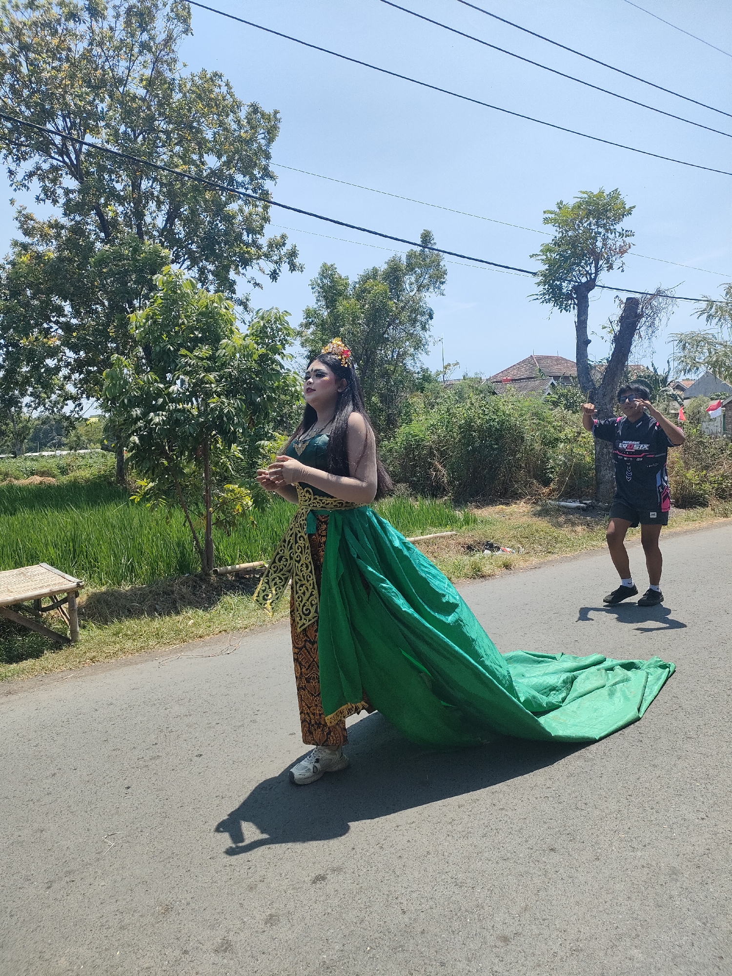 In Tongaswetan, Indonesia, on a bright, sunny day, two individuals are present on a paved road. A woman, adorned in a traditional green and gold outfit with a long, flowing green train, stands facing left. She has dark hair styled with flowers, dramatic makeup, and her hands are clasped in front of her. She is wearing white sneakers. To her right and slightly behind her, a young man in a black t-shirt and dark shorts is captured mid-motion, appearing to gesture with his hands and point his left arm upward, with his right hand near his face. He is also wearing dark sneakers. The background features lush green rice paddies, trees, and residential buildings. Overhead, power lines are visible against a clear blue sky. A small, woven bench sits on the left side of the road near the rice paddies. The scene appears to be outdoors, possibly during a cultural event or performance.