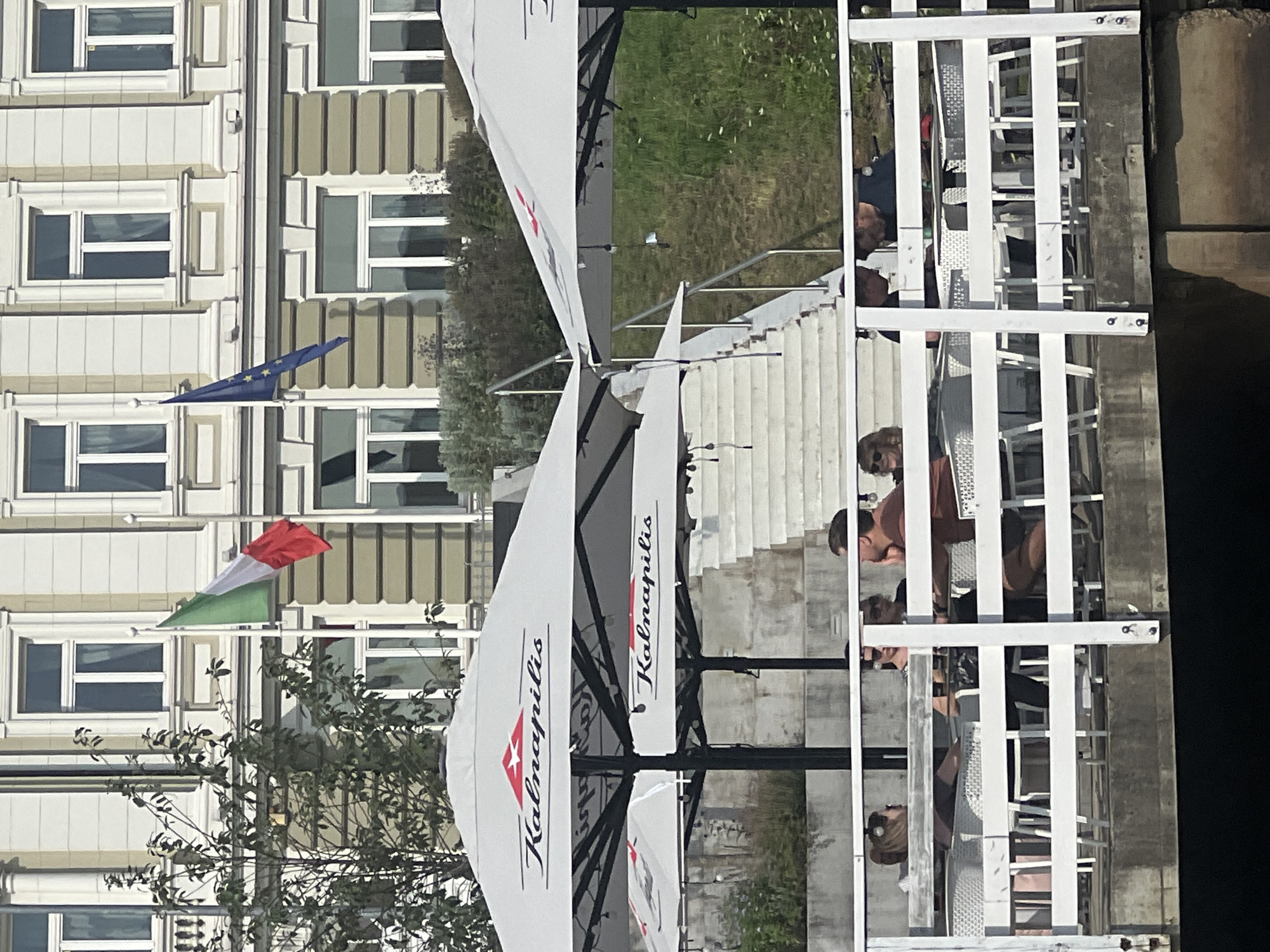 The image depicts an outdoor seating area of a cafe or restaurant in Vilnius, Lithuania, situated along a waterfront. Two large white umbrellas, branded with "Kalnapilis", are set up, providing shade. In the background, a light-colored building with multiple windows is visible, and two flags are flying: an Italian flag and an EU flag. A white staircase leads up from the seating area to the building. Several people are seated at tables on the white-railed deck, with some engaging in conversation and others looking at their phones. The overall scene suggests a relaxed, daytime atmosphere at a waterside establishment.