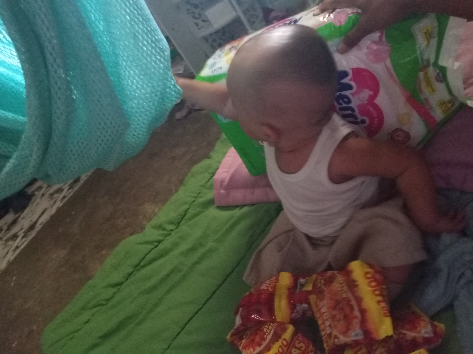 The image shows a baby sitting on a green padded surface, next to a stack of brightly colored snack packages. The baby is wearing a white tank top and brown shorts. To the left of the baby is a large piece of light blue netting. In the background, a person's hand is visible, reaching towards a package of diapers. The environment appears to be indoors, possibly a home. The lighting is dim, and the overall scene is slightly blurry.