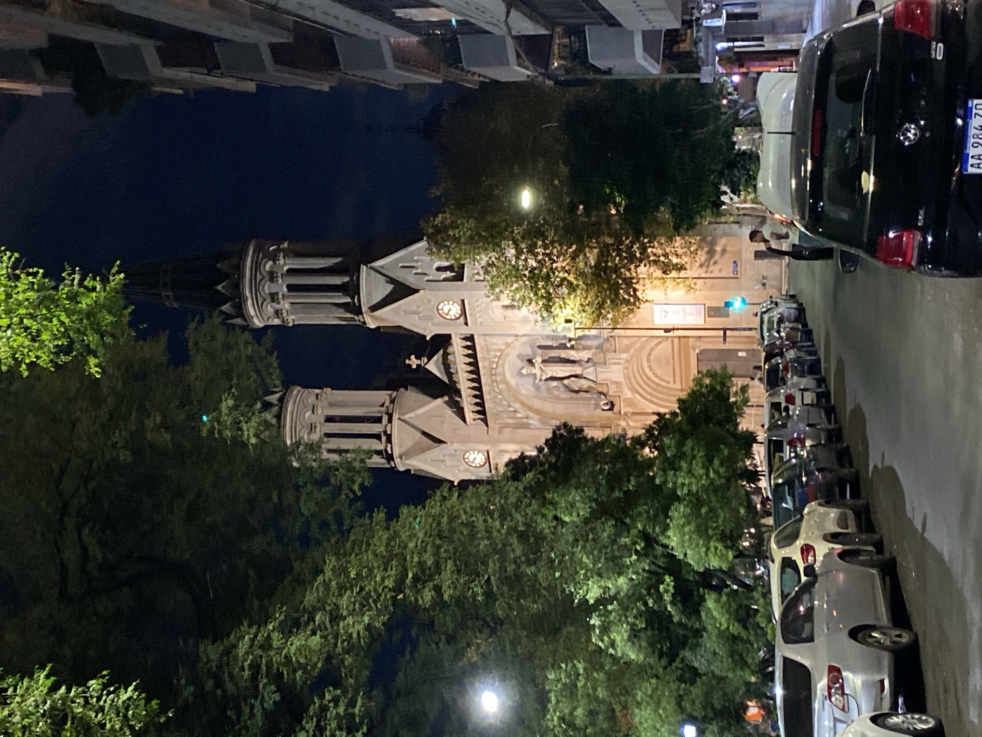 The image depicts the Metropolitan Cathedral of Buenos Aires, Argentina, at night. The cathedral is brightly illuminated. Trees obscure parts of the building. Cars are parked along the street to the right of the cathedral. One person appears to be standing near a parked car. The sky is dark.
