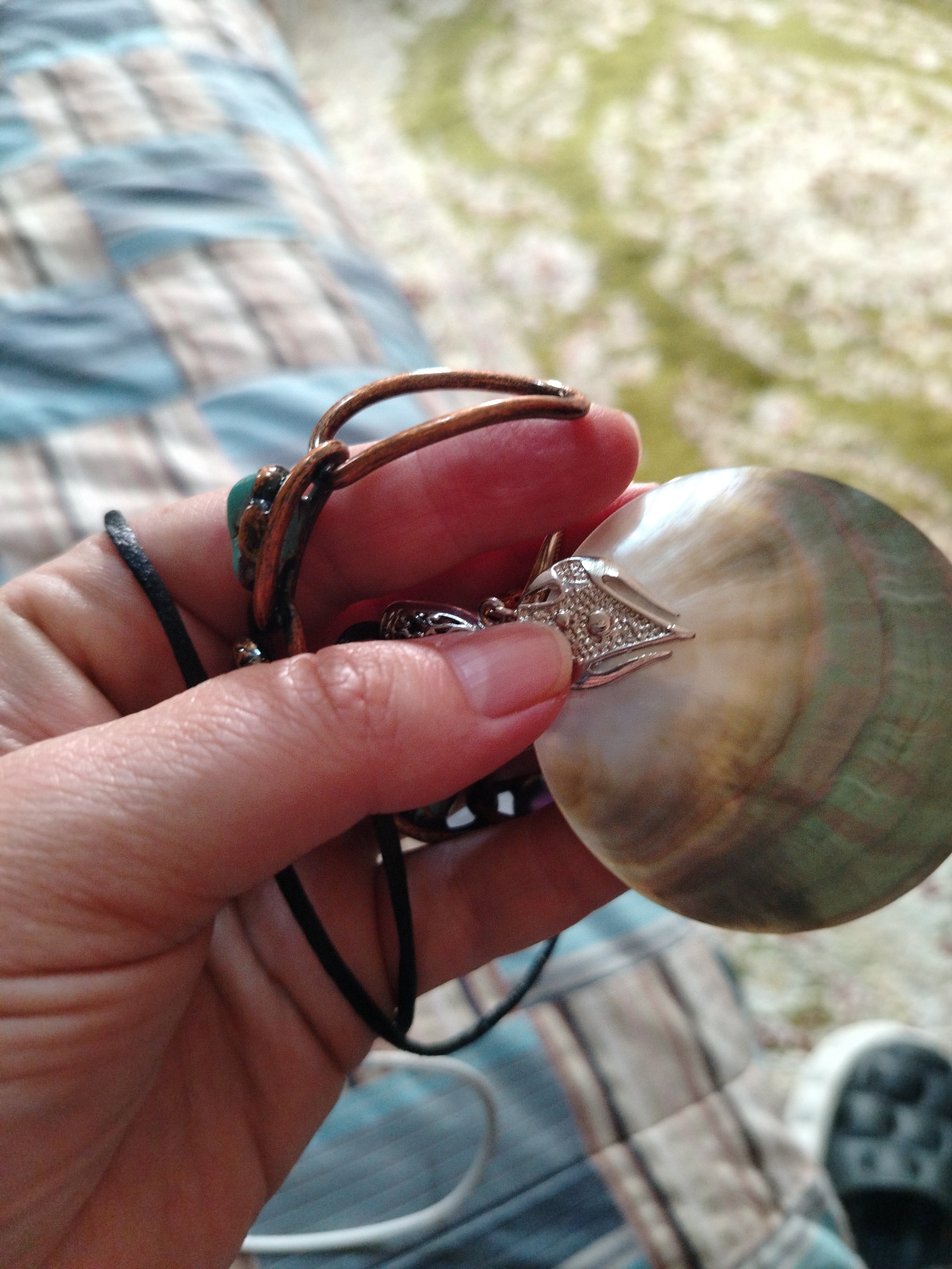 A person holds a necklace with a large, iridescent, circular pendant featuring a jeweled clasp. The necklace has a brown cord with turquoise accents and a black cord. The person's hand is visible, and a patterned bedspread and part of a shoe can be seen in the background. The location is not identifiable as Al Mas'oudi, United Arab Emirates, based on the visual information provided.