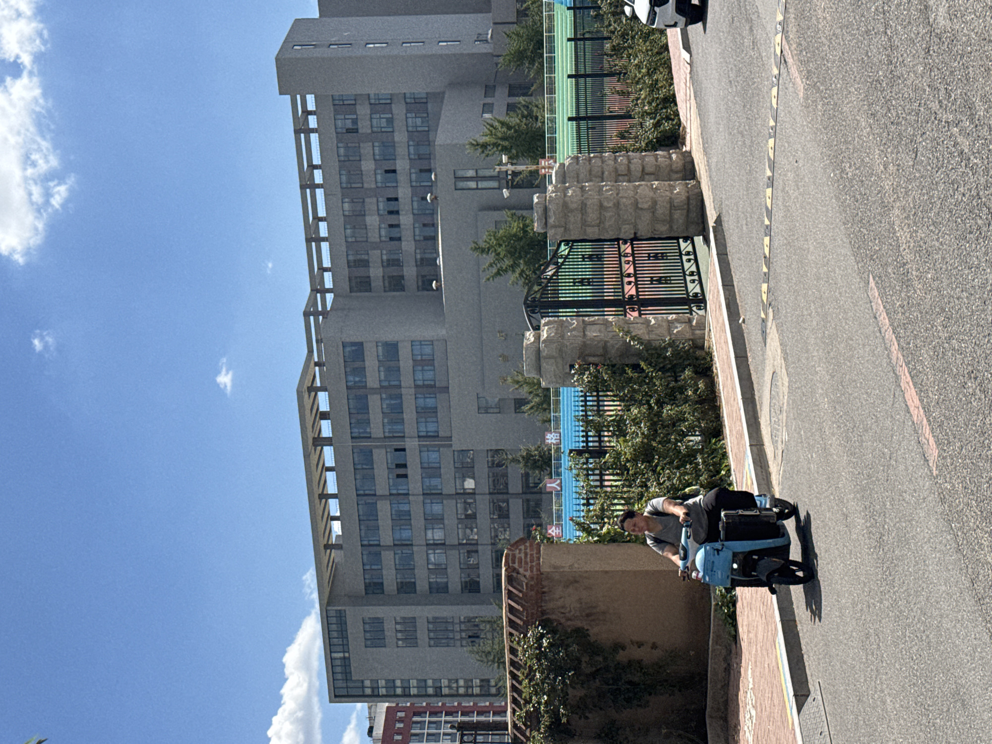 A medium shot captures a person riding a blue electric scooter on a paved road in Zhongtun, China. The rider, wearing a gray t-shirt and dark pants, is looking forward. The scooter has a large black storage container on the back. In the background, a large, grey, multi-story building with numerous windows stands under a blue sky with scattered white clouds. To the left of the building, a red-brick structure is partially visible. A black metal gate with ornate details is situated in front of the building, flanked by stone pillars. Green bushes and trees are visible in front of the gate and extending along the property line. A speed bump is present on the road in the foreground.