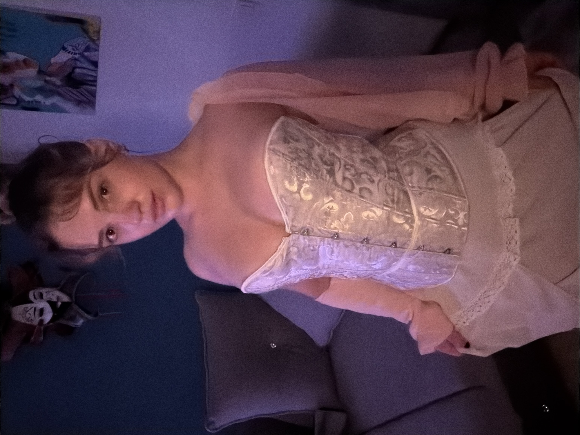 A young person, likely female, is captured in a vertical-orientation photograph, posing in what appears to be an indoor setting. She has light brown hair, styled back, and looks directly at the camera with a neutral to slightly serious expression. Her head is tilted, giving the image a dynamic feel.
She is dressed in an elaborate white or light-colored corset-style top adorned with intricate, possibly silver-toned, patterns and featuring visible hook-and-eye closures down the front. Beneath the corset, a flowing light-colored skirt or dress with delicate lace trim is visible. She wears light pink, fingerless arm warmers or long sleeves that extend up her arms, adding to the ensemble. Her fingernails are painted dark.
The background suggests a dimly lit room, possibly a bedroom or living area, with a soft purple and blue ambient light that indicates evening or night. To the left, a dark blue wall features a framed piece of artwork with abstract blue and white elements. Further left, near the bottom, a collection of decorative masks in white, black, and red are arranged on a dark surface. To the right, a lighter purple wall is visible. Below, a dark gray sofa with a cushion, featuring a small shiny embellishment, provides seating.
The image conveys a sense of quiet posing, emphasizing the subject's attire and gaze within a softly illuminated, almost theatrical atmosphere. While the provided context places this scene in Большая Шильна, Russia, there are no specific visual cues within the image itself to confirm this location. No visible text is present in the media.