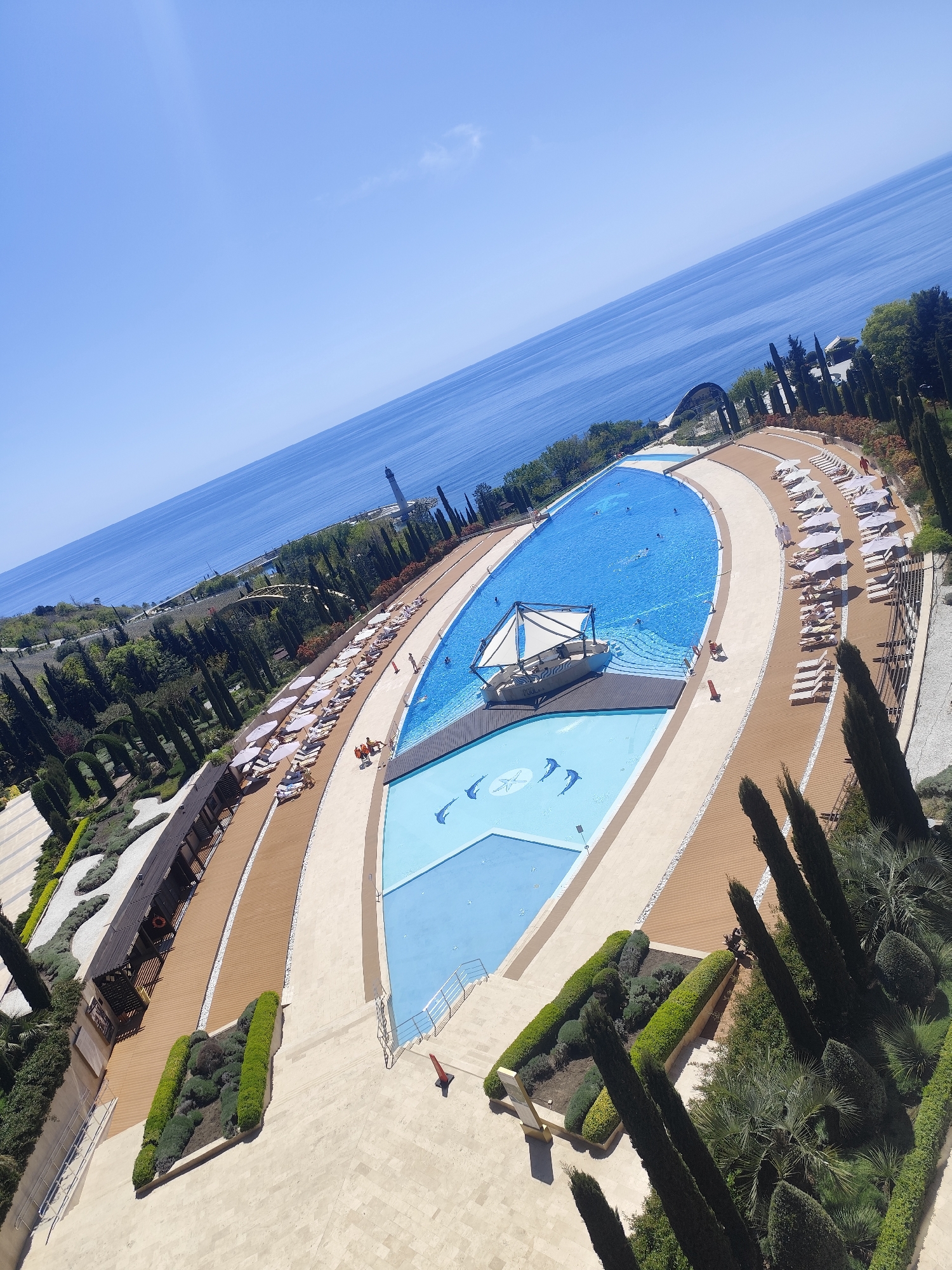 This high-angle, slightly tilted photograph captures an expansive, luxurious resort-style swimming pool complex set against a stunning backdrop of the deep blue sea. The sky above is clear and bright, indicative of a sunny mid-day.
The main subject is a large, irregularly shaped swimming pool, filled with several people swimming and enjoying the water. Within this pool is a distinctive boat-like structure, serving as a shaded lounge area with a white canopy, where the word "AQUA" is faintly visible. Adjacent to the main pool is a shallower section, decorated with playful dolphin patterns on its light blue floor.
Surrounding the pools are extensive paved and wooden deck areas, lined with numerous white sun loungers and matching umbrellas, some occupied by sunbathers. Pathways wind through meticulously manicured gardens featuring lush green foliage, including tall, slender cypress-like trees and decorative hedges. Small buildings or cabanas are integrated into the landscaping.
In the distance, the clear blue sea stretches to the horizon, with a noticeable lighthouse-like structure visible on the far coastline. The overall scene depicts a vibrant leisure environment, ideal for relaxation and recreation, suggesting a high-end coastal vacation destination.