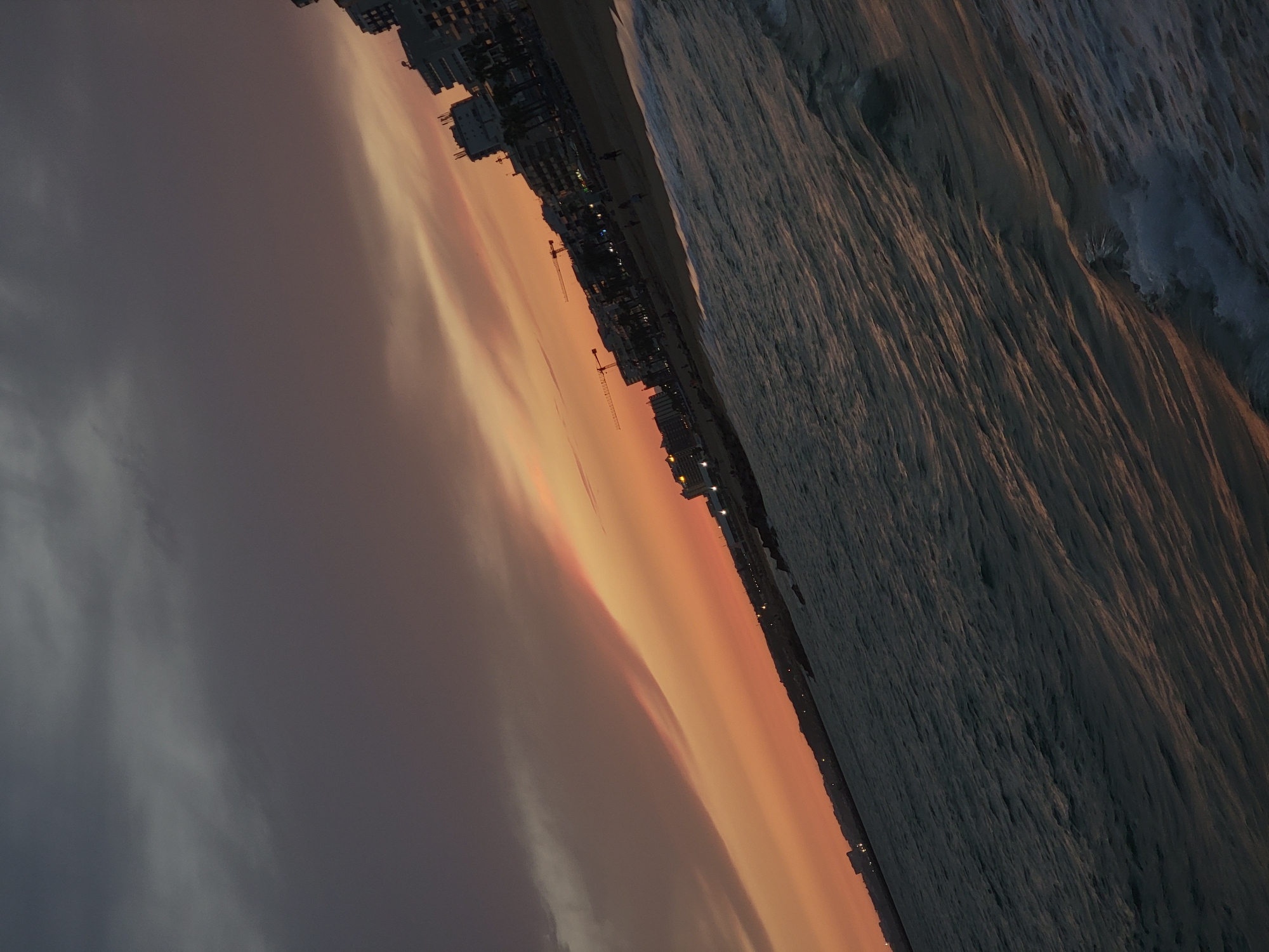 The image depicts a tilted perspective of a coastal scene at dusk, likely in Quarteira, Portugal, given the architectural style of the buildings. In the foreground, the dark, rippling water of the ocean is prominent, with faint reflections of the sky visible on its surface. The waves appear calm, with small white crests. Along the shoreline, a sandy beach is visible, sparsely populated with a few indistinct figures. Behind the beach, a row of multi-story buildings with a resort-like appearance lines the coast. Several construction cranes are visible amongst the buildings, indicating ongoing development. The sky above is a gradient of deep grays and purples near the top, transitioning to a soft orange and pink glow near the horizon, suggesting the sun has recently set. The overall lighting is dim, characteristic of twilight.