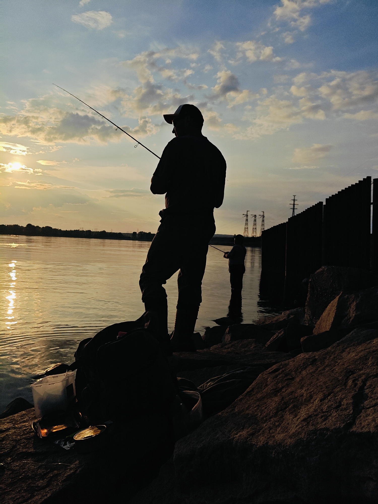 The image depicts two individuals fishing on a rocky shore at sunset. The foreground is dominated by a large, dark silhouette of a person wearing a baseball cap and fishing gear, with a backpack and fishing bucket to their left. The person is standing on a rocky embankment, with their fishing rod extended towards the water. The water reflects the warm glow of the setting sun, creating shimmering patterns on its surface. In the background, a second, smaller silhouette of a person is also fishing by the water's edge. Beyond the water, a treeline is visible, and further in the distance, industrial structures and power lines punctuate the horizon. The sky is a blend of blue and orange hues, with scattered clouds. The overall atmosphere is tranquil and serene. The scene is identifiable as being in the city of Chaikovsky, Russia, likely along the Kama River.