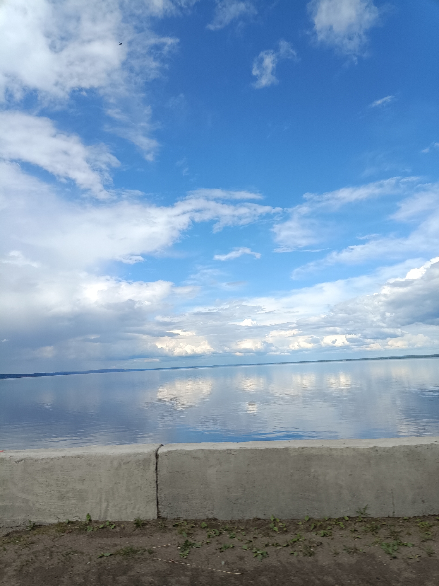 The image depicts a serene waterfront scene in Chaikovsky, Russia. A gray concrete wall, slightly cracked, runs horizontally across the lower portion of the frame, with sparse vegetation growing along its base. Above the wall, a calm body of water reflects the sky, displaying varying shades of blue and the shapes of puffy white clouds. The sky itself occupies the upper two-thirds of the image, filled with the same mix of blue sky and white, billowy clouds. A small, dark object, likely a bird, is visible in the upper portion of the sky. The overall scene is tranquil, suggesting a calm day along the waterfront.