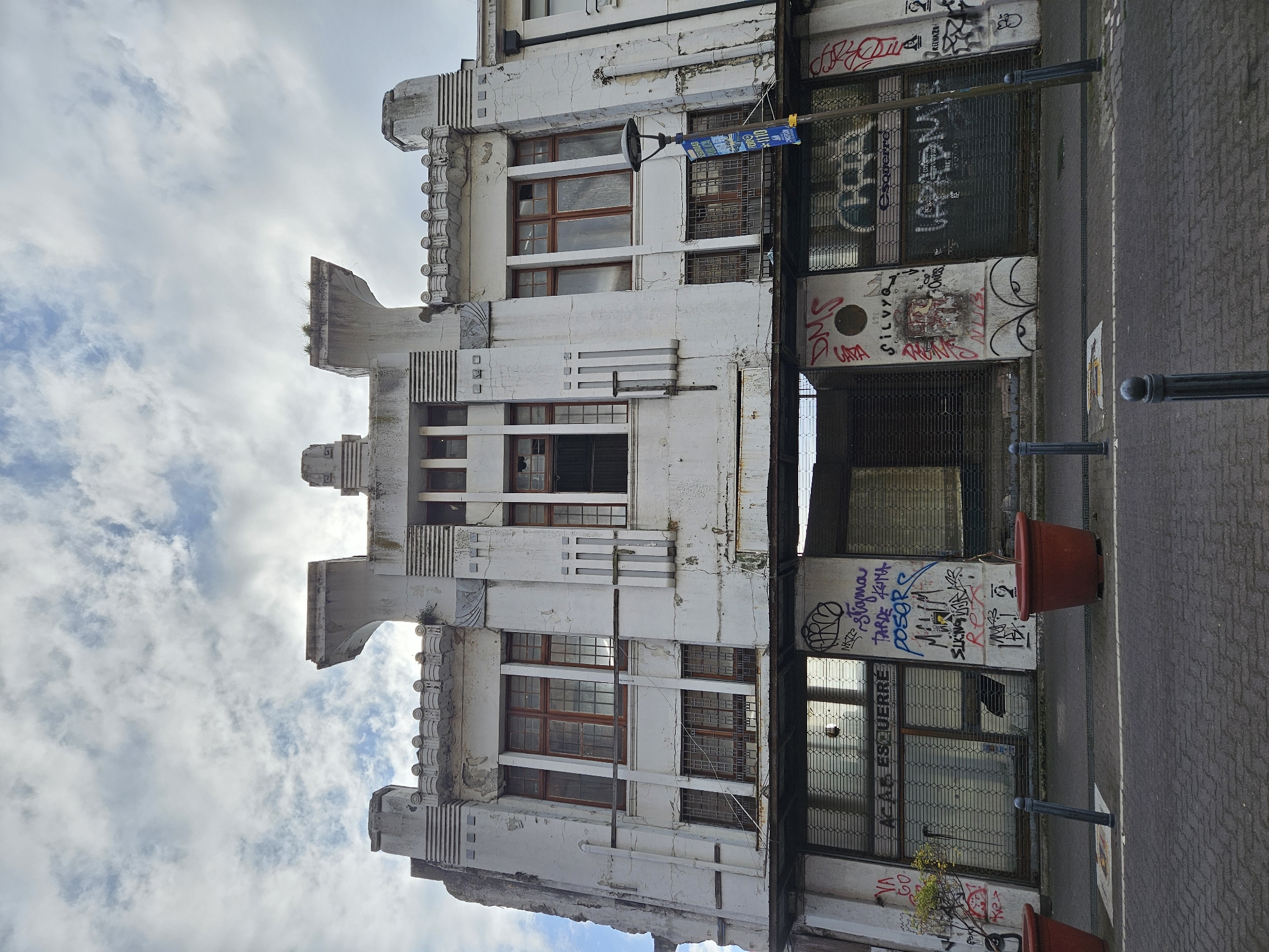The image showcases the facade of a distressed, multi-story building in Concepcion, Chile. Its architecture features an Art Deco style with prominent geometric shapes and tiered structures. The building's white exterior is heavily weathered and marked with graffiti. The windows are rectangular with visible wooden frames, some are boarded up, others show broken panes. Protective metal grills cover the storefronts, further defaced with graffiti. The adjacent street has a brick surface, adorned with two large terracotta plant pots flanking a streetlight, and another streetlight pole, all positioned in close proximity to the building. The sky visible above the structure is partly obscured by clouds.