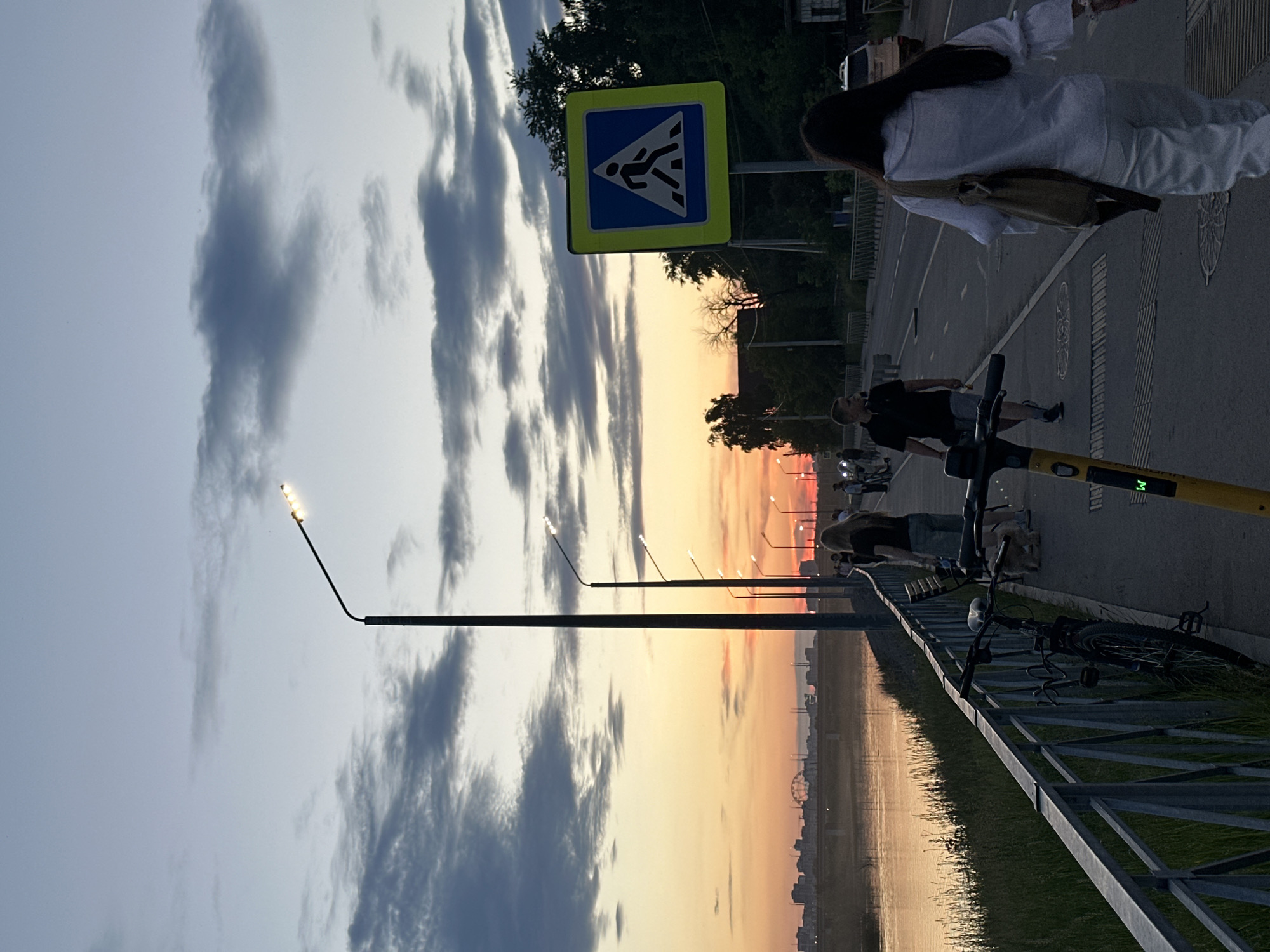 This media file captures an outdoor scene during sunset, likely in Irkutsk, Russia, given the context. The sky displays a gradient of colors from pale blue with dark, wispy clouds to vibrant orange and yellow hues as the sun sets, reflecting on the water in the foreground. A metallic fence runs along the edge of the water, with grass on the other side.
Several people are visible. In the background, a woman with long dark hair is walking away from the camera, wearing a white top and pants, and carrying a bag. Closer to the foreground, a man in a dark t-shirt and shorts is riding a scooter, and another man is walking. There are also glimpses of other people and vehicles, suggesting a public space.
Several objects are present. A yellow electric scooter with green lettering is prominently displayed in the foreground. A bicycle is parked near the fence. Streetlights with multiple lamps are lined up along the pathway, with some illuminated, indicating the approaching evening. A pedestrian crossing sign, a blue square with a white pictogram of a person crossing, is also visible, mounted on a pole. The scene appears to be a promenade or waterfront area, possibly a park or embankment, with a road running alongside. The overall mood is tranquil and atmospheric, characteristic of a sunset setting. No other visible text is decipherable.
