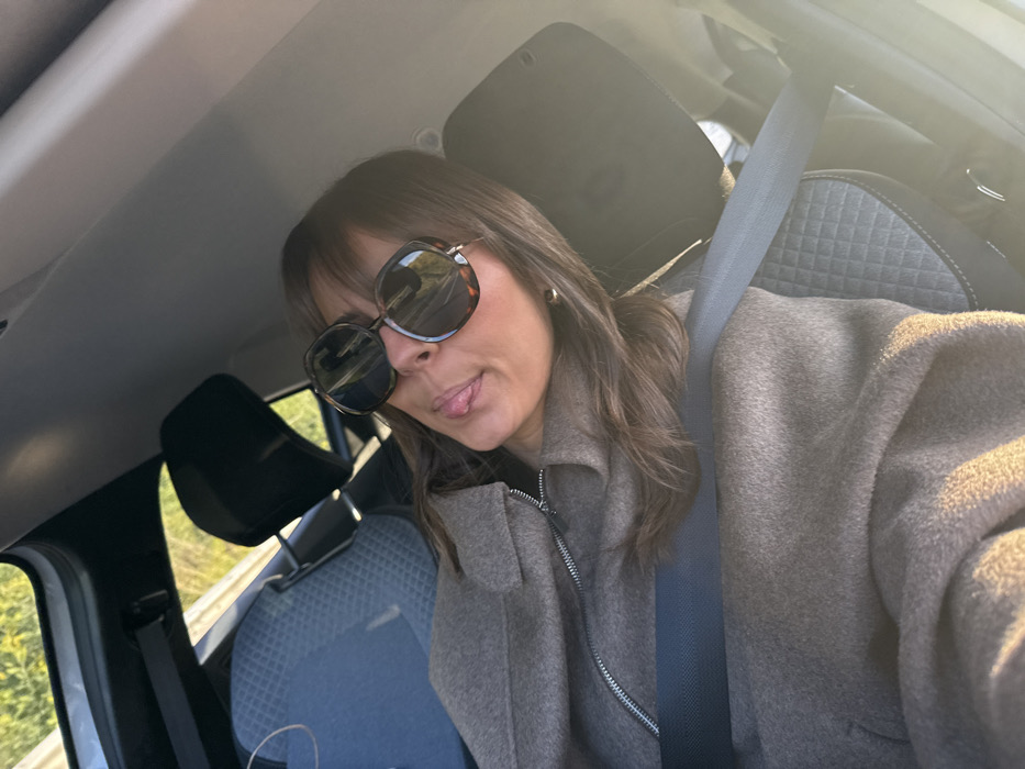 A woman is captured in a selfie taken inside a car. She is positioned in the driver's or front passenger's seat, with a light-colored car ceiling visible above her and dark headrests behind her. She has medium-length, dark brown hair with bangs and is wearing oversized, dark-rimmed sunglasses, possibly with a tortoiseshell pattern. Her head is tilted slightly, and she is playfully sticking out her tongue. A small, gold-colored hoop earring is visible on her right earlobe. She is dressed in a thick, light brown or taupe-colored coat with a prominent silver zipper down the front, indicating a warm, wool-like material. A light grey seatbelt is fastened across her chest.
The car's interior features grey seats with a textured, possibly quilted, diamond pattern visible on the headrests and seat upholstery. Through the car windows on the left side of the image, green foliage and what appears to be a road barrier or railing can be seen, suggesting the car is either parked by a road or in motion. The overall lighting is bright and sunny, casting a warm glow on her coat and creating reflections on her sunglasses, indicating daytime, likely morning or late afternoon due to the angle of the sunlight. No text is visible in the image.
