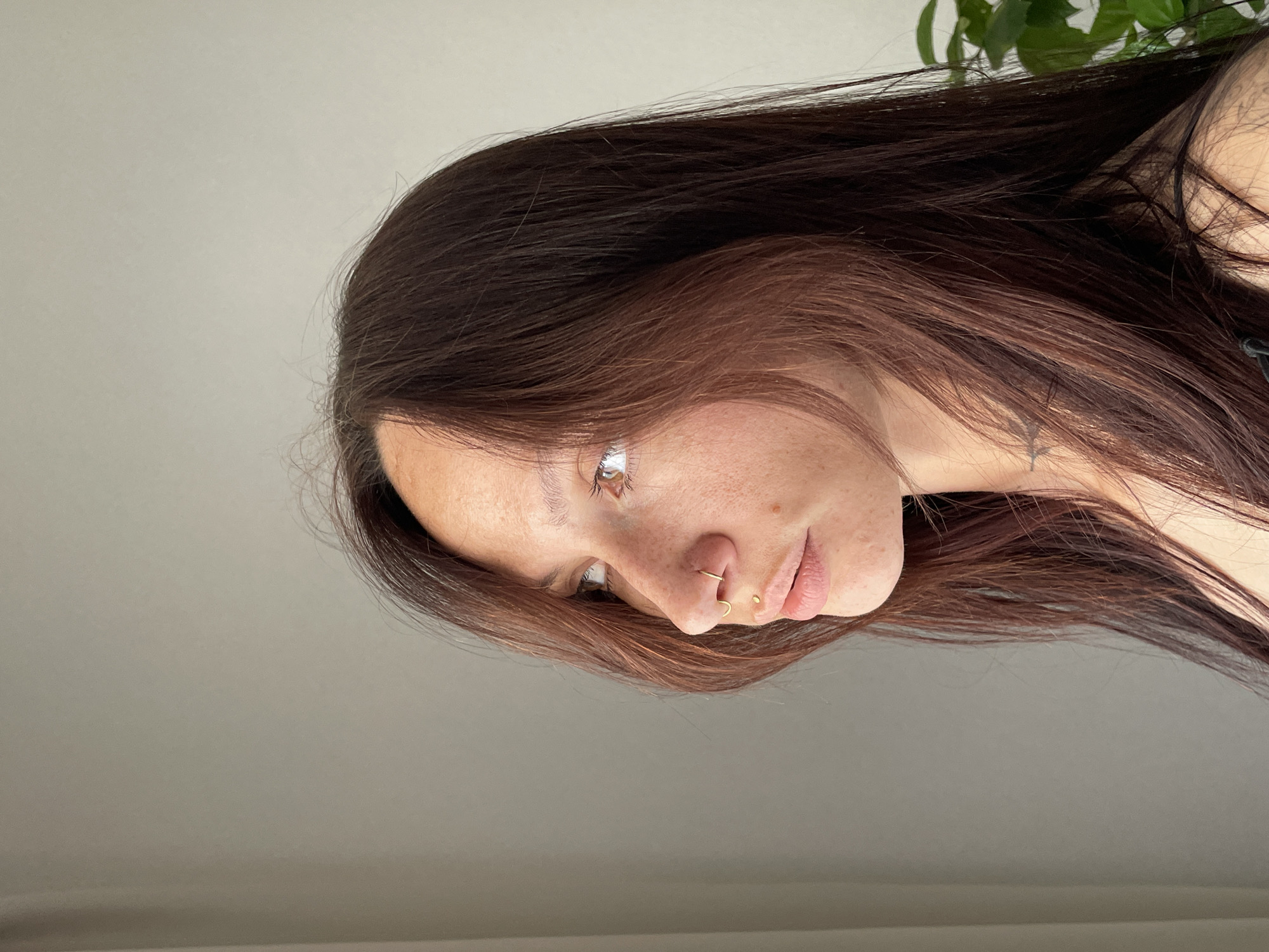 The image shows a close-up profile of a young Caucasian woman with long, dark auburn hair. She has fair skin with freckles, light brown eyes, and is looking to the left. She is wearing gold jewelry, including a septum ring and a small hoop in her nostril, as well as a labret piercing. Her lips are slightly parted, and her expression is neutral. Her shoulders and upper chest are visible, and she appears to have tattoos on her neck and shoulder. The background is a plain, light gray wall. The lighting suggests an indoor setting, possibly near a window due to the slight highlighting on her face. There is a hint of a green plant in the lower right corner. The image does not contain any discernible elements that would allow for the identification of a specific location in Pervomaysky, Russia.