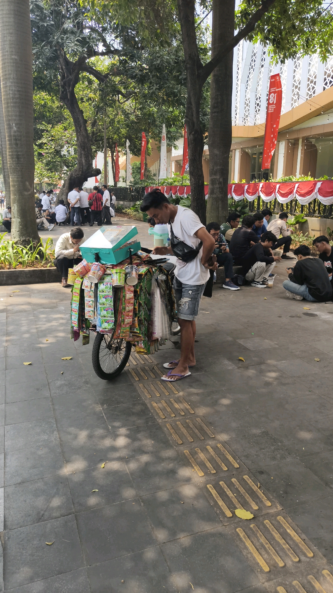A young man in a white t-shirt and denim shorts is standing next to a mobile cart filled with snacks and drinks. He is looking down at the cart and appears to be arranging or selecting items. The cart has a teal-colored top with transparent compartments and is decorated with numerous small packets of food items and drinks. The man is wearing purple flip-flops.
In the background, a group of people are sitting on the ground and on a low wall, some of whom are looking at their phones. Behind them, there is a building with a modern facade decorated with white latticework and red banners featuring the number "81" and text in Indonesian. The scene takes place outdoors on a paved walkway with tactile paving for the visually impaired. Trees with lush green foliage provide shade, casting dappled sunlight on the ground. The overall setting suggests a public space in Central Jakarta, Indonesia, possibly near a university or public gathering area, given the presence of many young people and the celebratory banners. The red and white decoration on the building and banners indicates a national event, likely the Indonesian Independence Day celebrations, which occur in August.