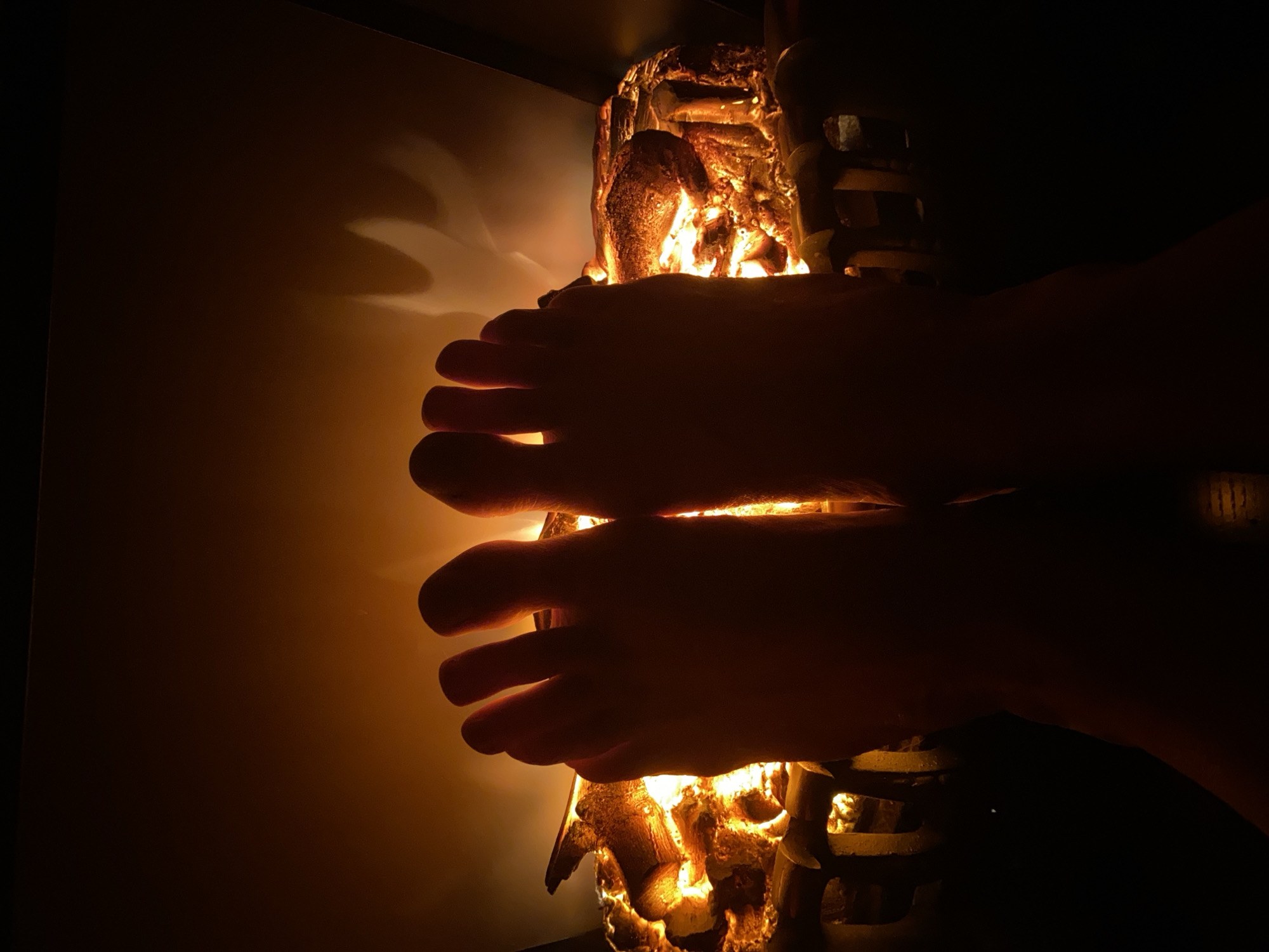 The image shows a pair of feet held against a light source, which appears to be a decorative lamp. The lamp has an organic shape and casts an orange light. The wall behind the lamp is illuminated by the light, while the surrounding areas are dark. The scene is dimly lit, focusing on the interplay of light and shadow. There is no indication that the photo was taken in Novokuznetsk, Russia.
