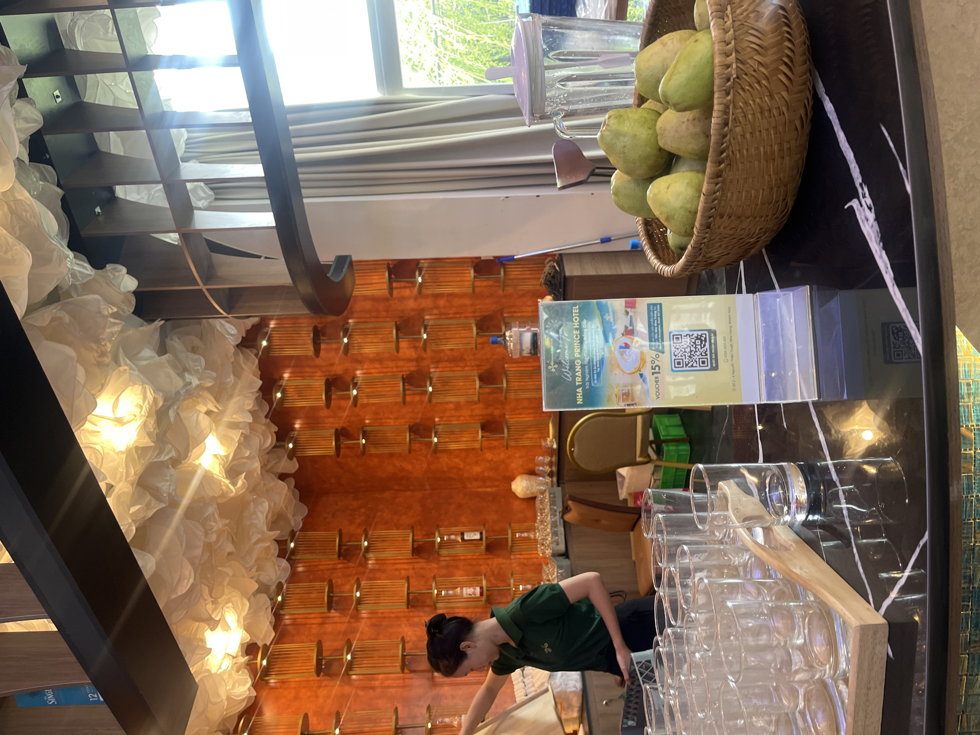 In an indoor establishment, likely a hotel lobby or buffet area, a woman in a green polo shirt stands at a counter. She is partially visible from the side and is holding a white cloth. In front of her, on the dark, reflective counter, are multiple rows of clear drinking glasses. A wooden serving tong rests on the glasses. To the right of the glasses, a sign with text and a QR code is displayed. Beyond the sign, a woven basket is filled with several guavas. Behind the basket, a large clear pitcher with a light purple lid stands on the counter. The background features an orange wall adorned with vertical wooden slats and decorative elements. Above, a ceiling is decorated with white, cloud-like fabric illuminated by warm lights. Further back, a tall shelving unit with curved edges is visible, partially obscured by beige curtains and a window.