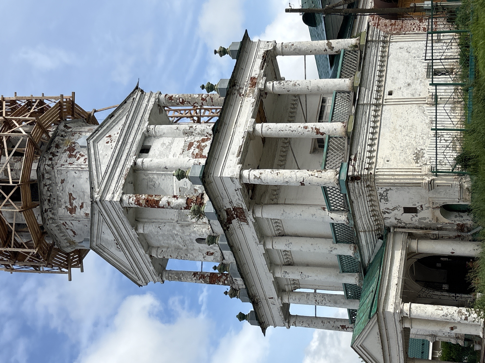 The image shows the exterior of the Church of the Transfiguration in Kstininio, Russia, under repair. The church is a white, multi-tiered structure with classical architectural elements, including columns and pediments. The paint is peeling, revealing brickwork underneath, indicating age and a need for restoration. Scaffolding is visible at the top of the structure, suggesting ongoing construction or repair work. The sky is blue with scattered clouds. The lower part of the church has a green roofed entrance with columns. A green metal fence with a diamond pattern is in the foreground, with tall green grass growing behind it. No people are visible in the image.