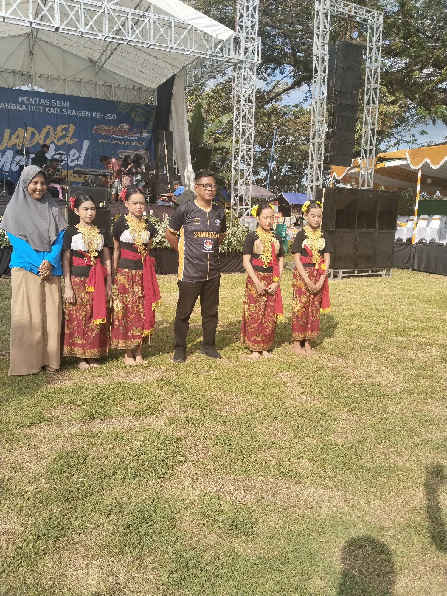 In Sambirejo, Indonesia, a group of six individuals stands on a grassy field in front of a stage structure. Four of the figures are barefoot, young women dressed in traditional black and red attire. The two women flanking the young dancers are wearing everyday clothes and headdress. The background reveals a stage set up for a performance. The stage backdrop bears the words "PENTAS SENI," "ANGKA HUT KAB. SRAGEN KE-280," and "JADDEL Martel." A sound system with speakers is set to the side of the stage. The shadow of an individual in the lower right corner of the image, suggests a photographer.