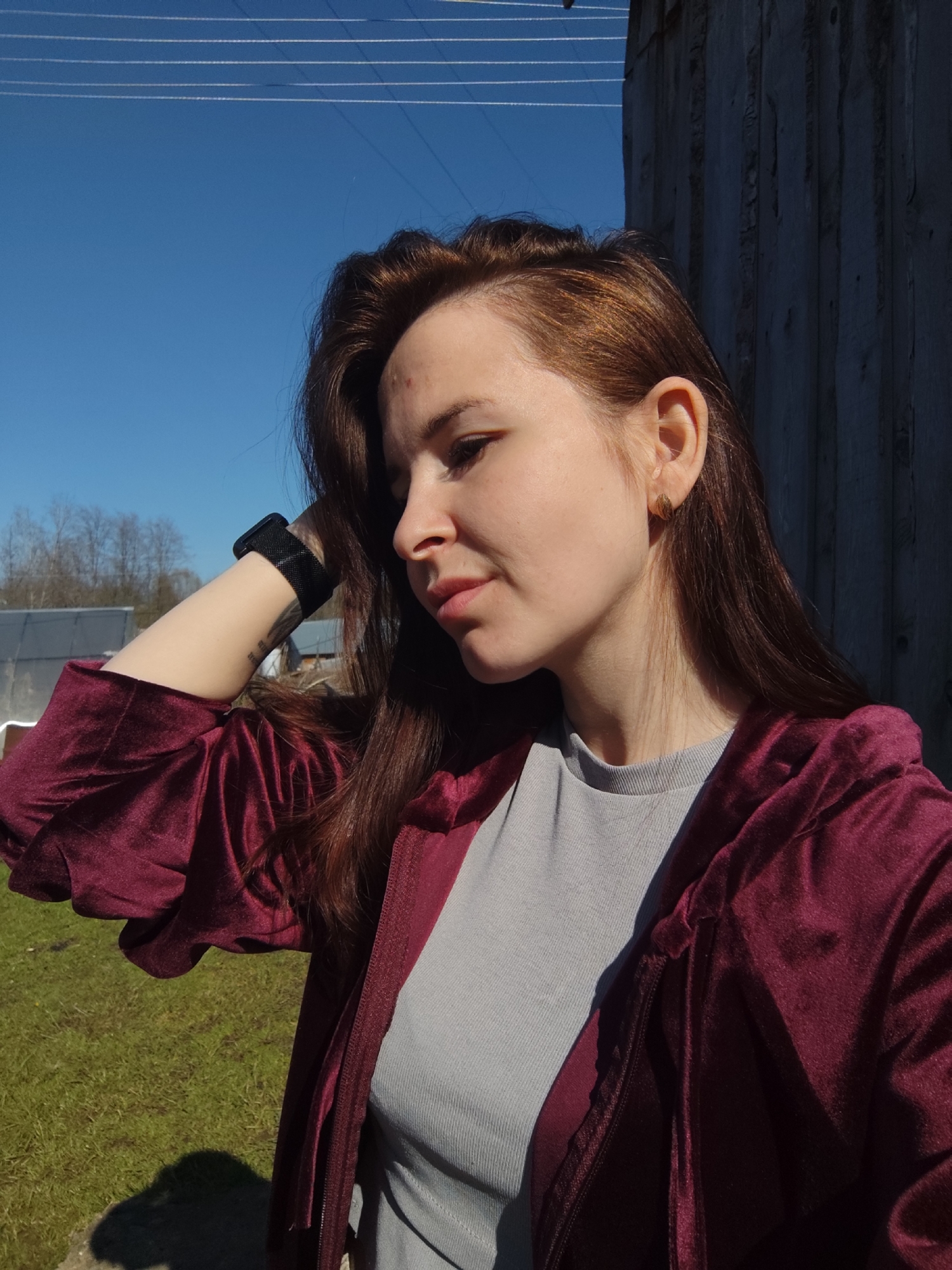 A young woman with auburn hair is outdoors, standing near a wooden building. She wears a gray top and a dark red velvet jacket. Her left arm is raised, with her hand resting on her head, showcasing a black smartwatch. The setting is a sunny day with a clear blue sky. The background includes trees and a building with a light-colored roof. This location is in Фоки, Russia.
