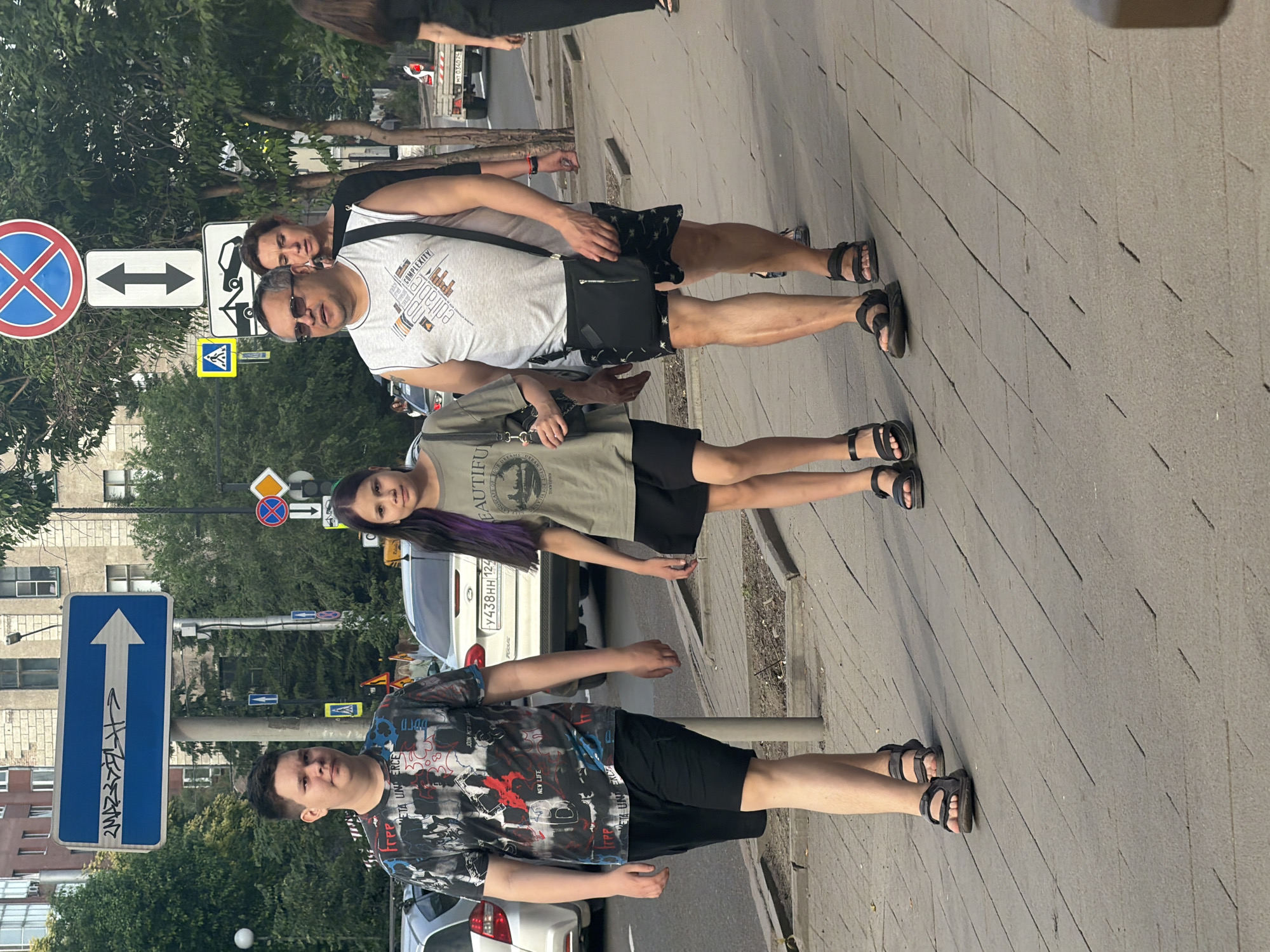 Four people, two adults and two children, walk along a sidewalk in Krasnoyarsk, Russia. The adult male on the right wears a grey tank top and black patterned shorts, carrying a black crossbody bag. He is accompanied by a woman in a black top. To their left, a girl with purple-streaked hair wears a green t-shirt and black shorts. To the far left, a boy in a graphic t-shirt and black shorts walks ahead of the group. They are on a paved sidewalk next to a street with cars and traffic signs. The background shows buildings and trees.