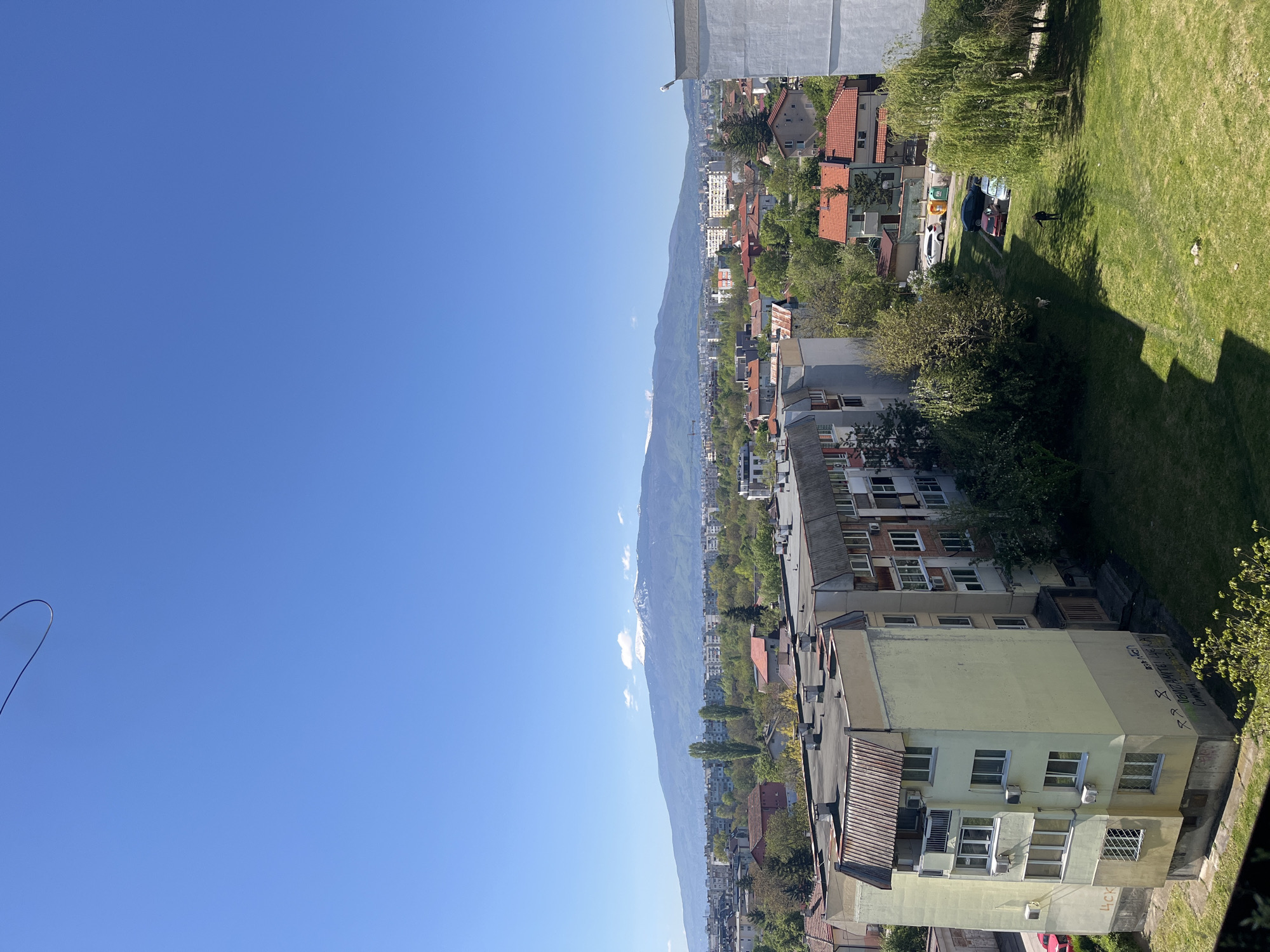 An eye-level, daytime shot looking down at a scene of Sofia, Bulgaria, on a sunny day. The sky is mostly clear blue. In the distance, a large mountain range is visible, running horizontally across the frame. Buildings, interspersed with trees, span from the mountain range towards the viewer. The buildings vary in height and color, with some having red-tiled roofs. A green grassy area is visible in the upper-right portion of the frame. The view extends towards the lower portion of the frame, showing residential buildings with multiple windows. The image is captured in an upright position with the horizon oriented along the x axis. In the upper right, a person with a dog walks on the green lawn. Some graffiti can be seen on the side of a building in the lower right.