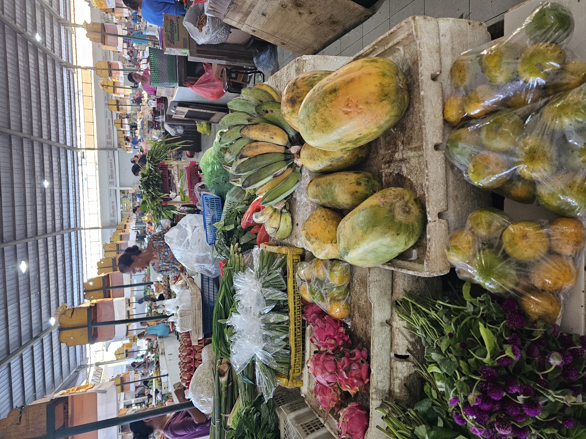 The image depicts a vibrant marketplace scene, likely in Denpasar, Indonesia, given the location mentioned. The foreground showcases a diverse assortment of produce on a vendor's stall: papayas, bananas, oranges in plastic bags, dragon fruit, spring onions, and leafy green vegetables. Purple globe amaranth flowers are also present. The stall is composed of a weathered concrete structure, typical of marketplaces. In the background, a bustling market setting is visible, with numerous vendors and customers under a large, corrugated metal roof supported by metal beams. A vendor is seen interacting with customers amid stacks of goods. The atmosphere is active and filled with the energy of commerce.