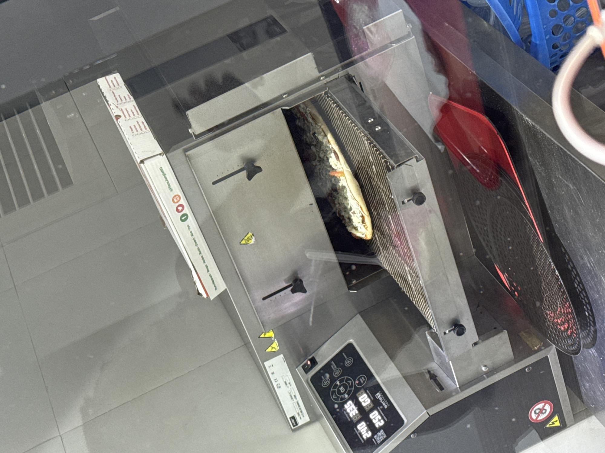 Here is a forensic description of the image:
The image appears to be taken indoors, possibly in a commercial kitchen. A pizza is visible inside of what looks like a conveyor pizza oven. The pizza is partially cooked and still has toppings on it. A pizza box is next to the pizza oven. The control panel indicates that the temperature setting is 250, potentially referring to degrees Celsius. Based on the text on the pizza box, it is located in Port-Harcourt, Brugu, Asaba, Banin, Oweni, Aba, and Uyo.