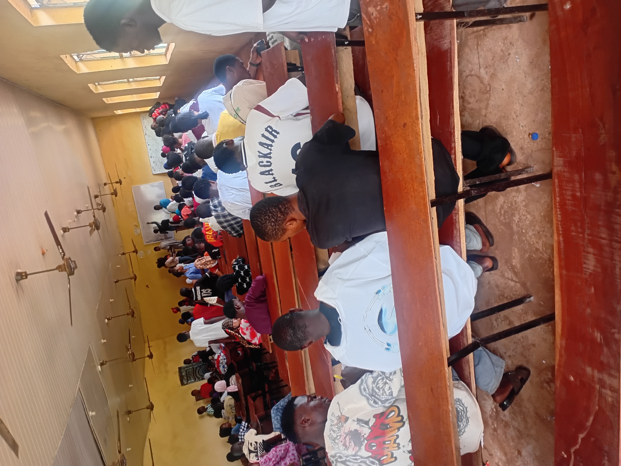 In Ogbogbo, Nigeria, a classroom scene unfolds with a group of young individuals, predominantly male, seated on wooden benches. The room appears to be a lecture hall, indicated by the presence of a whiteboard at the front and ceiling fans for ventilation. The students are casually dressed, with some wearing t-shirts featuring various graphics and text. One individual in the foreground wears a white t-shirt with the inscription "BLACKAIR." Many students are looking towards the front of the room or down at their laps, suggesting they are attentive to a presentation or lecture. The walls are painted a pale yellow, and the floor is a reddish-brown dirt. Sunlight streams in through windows on the right side of the room, illuminating the space. The overall atmosphere seems to be one of focused learning within a modest educational setting.