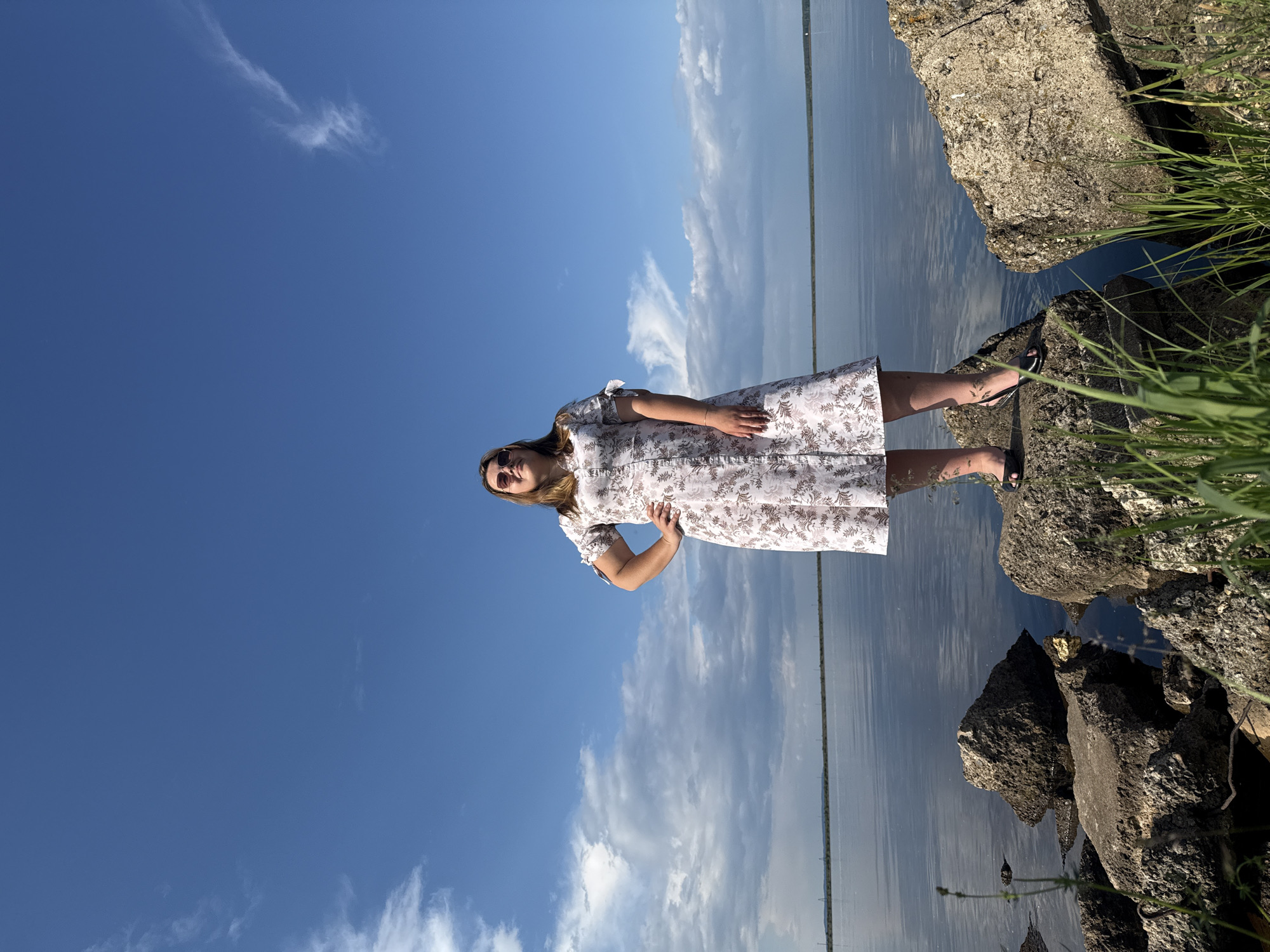 A young woman stands on large rocks by a body of water, her left hand on her hip, gazing upwards with a slight smile. She wears a light-colored, knee-length dress with a floral pattern and sunglasses. The sky above is a bright blue with scattered white clouds. The water is calm, reflecting the sky. The scene appears to be set on the shore of the Kama River in Chaikovsky, Russia, with a distant shoreline visible.