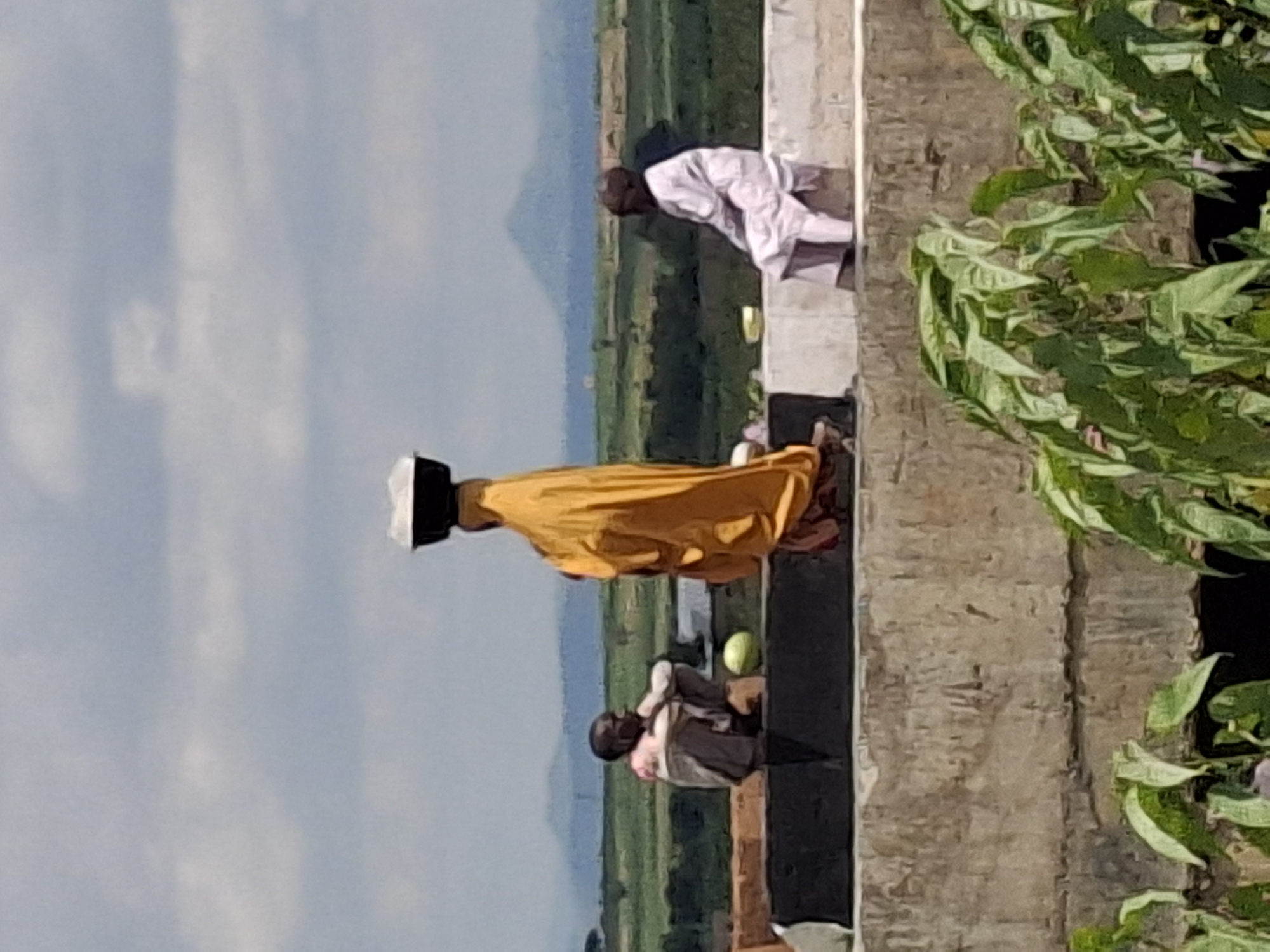 In Kumo, Nigeria, three individuals are visible outdoors. A person in the foreground on the left is squatting, facing right, wearing a light-colored shirt with pink accents and dark pants. Next to them, a person in the center stands with their back to the camera, draped in a bright yellow garment and wearing a dark bowl filled with white material on their head. To the right, another individual sits facing left, dressed in a light-colored robe. They are positioned on a raised concrete structure with a dark band along the top edge. Behind them, a green, hilly landscape stretches to the horizon under a cloudy sky. A large green object, possibly a watermelon, is visible on the ground between the first two individuals. In the far background, hazy mountains are discernible. Green foliage occupies the lower portion of the frame.