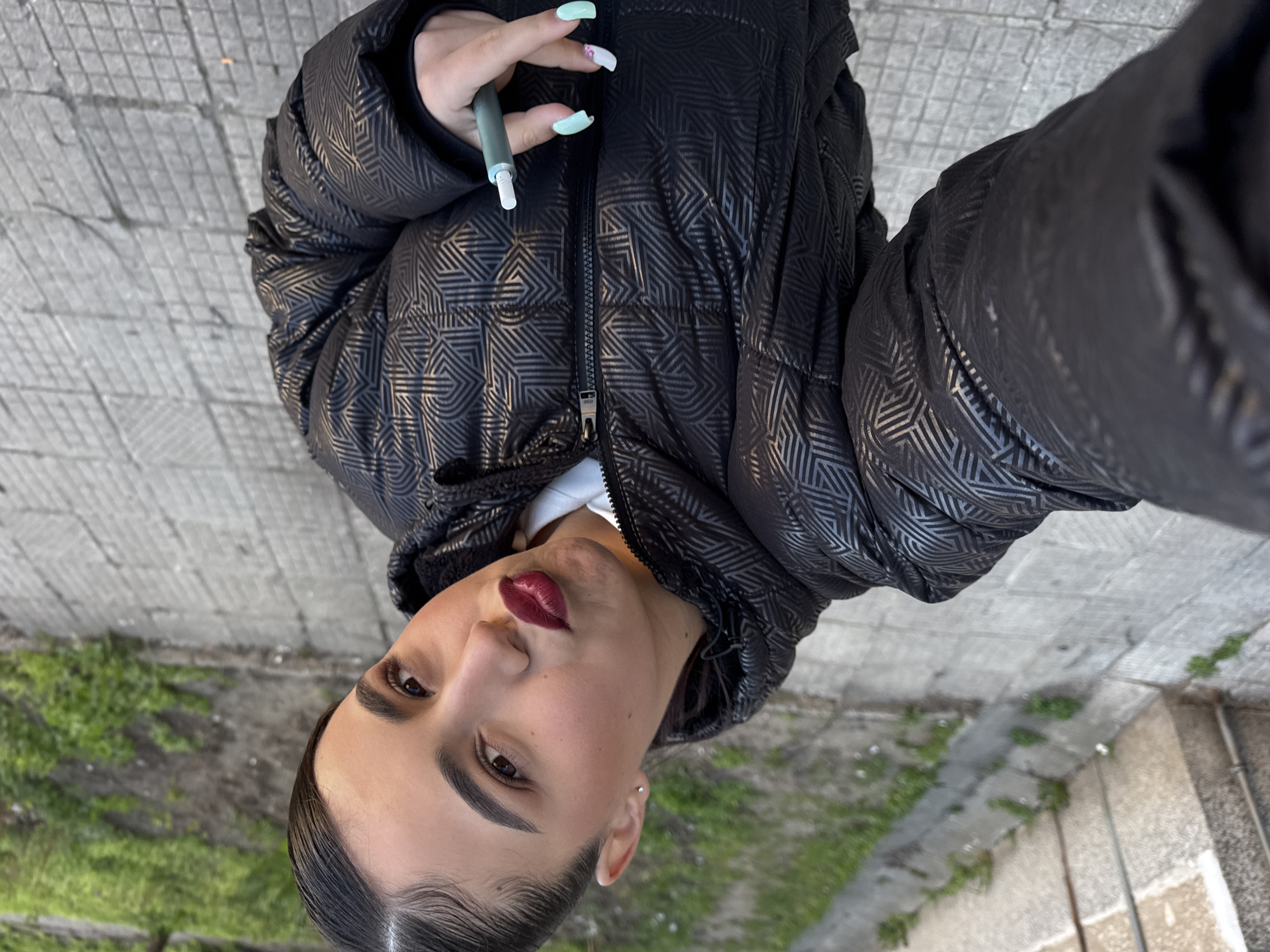 A young woman with slicked-back dark hair and dark red lipstick is holding an electronic cigarette in her right hand. She is wearing a black puffer jacket with a geometric pattern. The woman is standing outside, near what appears to be a paved surface with some greenery visible in the background. The image is taken from a slightly elevated angle, suggesting she might be holding the camera herself. Location cannot be accurately determined, Sofia, Bulgaria is not evident.