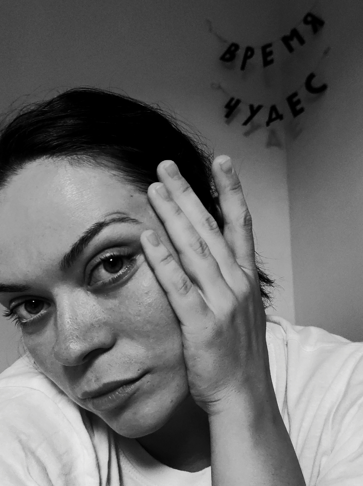 This black and white photograph presents a close-up portrait of a woman, looking directly into the camera. Her dark hair is styled away from her face. Her left hand is raised, with fingers gently splayed, resting against the left side of her face (viewer's right), partially obscuring her temple and cheek. Her eyes are prominent and dark, conveying a thoughtful or pensive expression, with her gaze directed forward. Her lips are slightly parted. She is wearing a light-colored, likely white, t-shirt, visible around her neckline and draped over her shoulder.
The background consists of a plain wall, rendered in shades of gray. In the upper right corner, a two-line string of letters is visible. The upper line reads "ВРЕМЯ" and the lower line "ЧУДЕС" in Cyrillic script. This translates from Russian to "Time of Miracles" or "Miracle Time." The lighting is soft, creating subtle shadows that add depth to her features and the texture of her skin. The overall mood is contemplative and intimate, enhanced by the monochrome treatment of the image. The setting appears to be indoors, possibly a personal space.