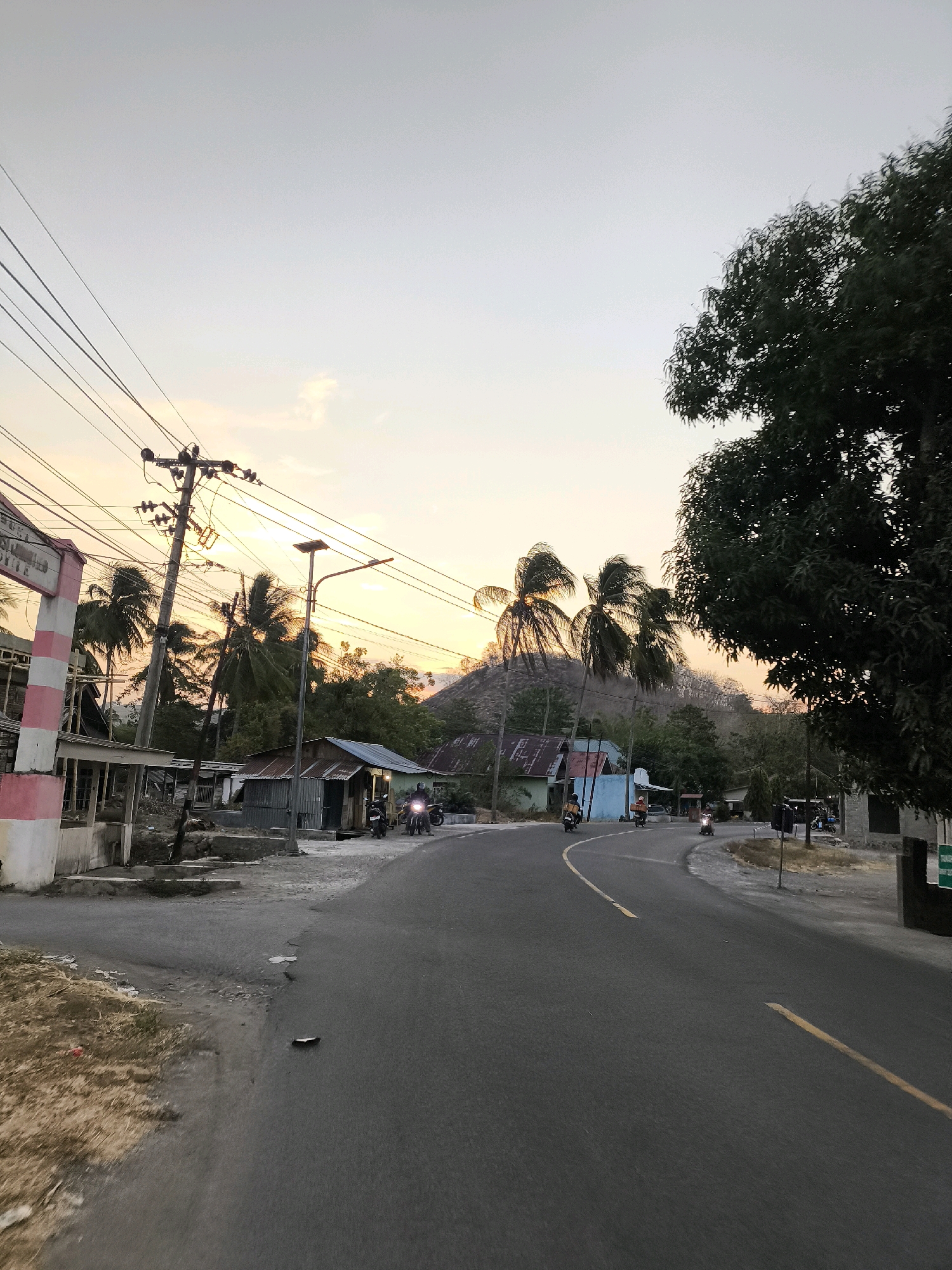 The image depicts a road scene at dusk in a rural area of Indonesia. A paved road with yellow lane markings curves through the frame, lined by houses and vegetation. Several motorcycles are present on the road, with some riders visible. Power lines stretch across the upper portion of the image, supported by poles. In the background, a rounded, tree-covered hill rises against a pale orange and yellow sky, indicating the setting sun. Coconut trees are scattered throughout the scene, and a large, leafy tree dominates the right side of the foreground. The environment appears to be a small village or settlement.