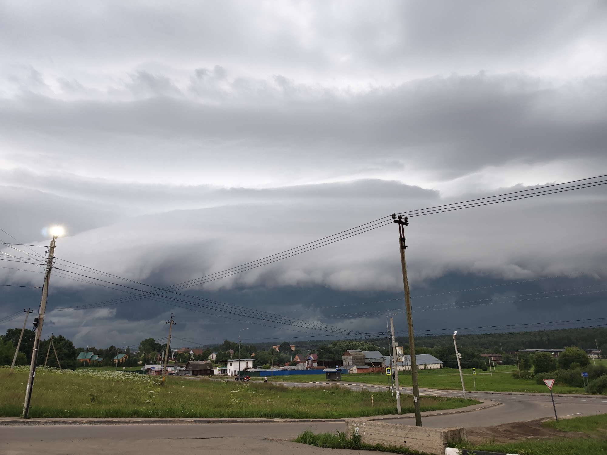 This image shows a rural scene in Ateptsevo, Russia, under a dramatic, stormy sky. The sky is filled with dark, ominous clouds that suggest an approaching storm, with a band of lighter, layered clouds stretched across the horizon. In the foreground, a paved road curves to the right, bordered by green grass and wildflowers. Several utility poles with wires are visible, including one with a lit lamp on the left. In the middle ground, a cluster of houses and other buildings forms a small village. Trees and greenery surround the village, with a dense forest forming the backdrop. A few signs, including a yield sign, are visible on the side of the road. There are no people or vehicles prominently featured in the image. The overall atmosphere is one of impending weather change in a quiet, rural setting.