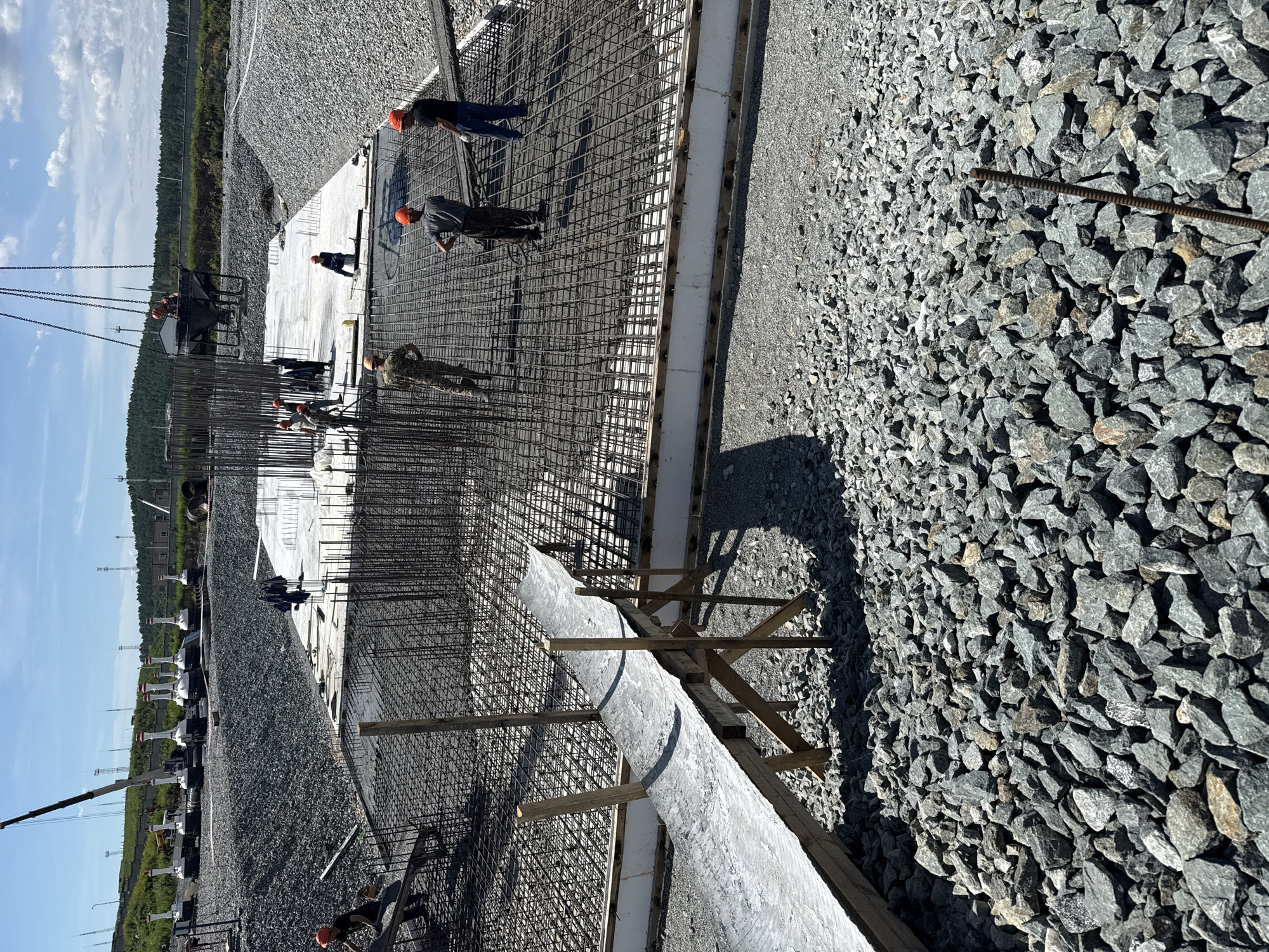 In an outdoor construction setting, several workers are engaged in pouring concrete onto a reinforced steel structure. The scene takes place on a clear, sunny day with a blue sky and scattered clouds. The ground is covered with a layer of grey gravel.

In the foreground, a large area of rebar is visible, forming a dense grid. A wooden ramp, covered with a grey material, leads up to this rebar structure. Several workers in hard hats and work clothes are present on and around the rebar. One worker, wearing an orange hard hat and a camouflage uniform, is walking on the rebar. Another worker, also in an orange hard hat, is standing near the edge of the rebar, observing the activity. To the right, two workers are in the process of pouring concrete; one is holding a hose, and the other is guiding the flow.

In the middle ground, a larger concrete slab is visible, also with rebar. A crane is positioned in the background, with a concrete bucket suspended from it, ready to be emptied. Several more workers are on this slab, some appearing to be directing the concrete flow.

Further in the background, a line of white and red industrial equipment, resembling large buoys on wheeled bases, is situated on a gravel embankment. Beyond them, a landscape of rolling hills covered in green vegetation stretches to the horizon, under the expansive sky. The presence of a construction site with heavy machinery, rebar, and concrete pouring indicates an ongoing construction project. The location is identified as the city of Leninsk, Russia.