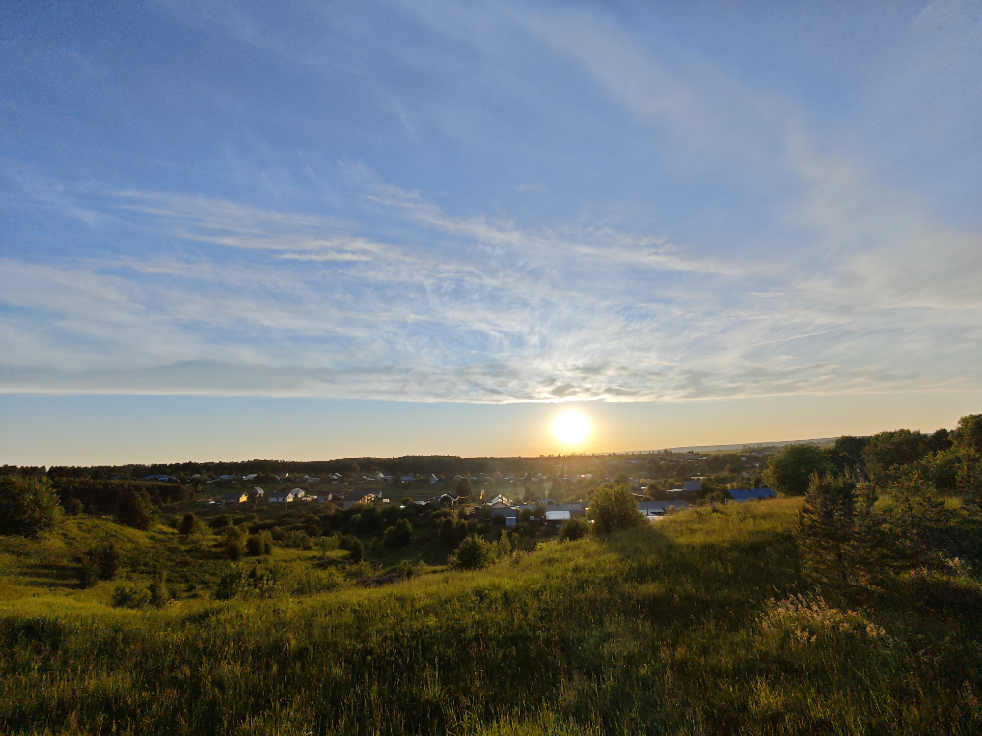 The image captures a serene rural landscape at sunset, looking down from a grassy hillside towards a village nestled in a valley. The bright, setting sun is positioned slightly to the right of the center horizon, casting a brilliant golden glow and creating a sunburst effect through thin, wispy clouds. The sky above is a clear, vibrant blue, gradually fading to a lighter yellow-orange closer to the horizon where the sun is located. Long, streaky white clouds stretch across the upper sky, particularly prominent on the left side.
In the midground, the small village consists of numerous houses with various roof colors, predominantly grey and blue, scattered amidst lush green trees and foliage. The warm light of the setting sun illuminates parts of the village, creating highlights on roofs and buildings, while other areas are beginning to fall into shadow. Beyond the village, a dense line of trees marks the distant horizon.
The foreground is dominated by a sun-drenched, rolling grassy hill, filled with tall, golden-green blades of grass and scattered wildflowers. Shadows are long and soft, extending from the unseen right side of the frame towards the left, indicating the low angle of the sun. No people or visible text are present in the image. The overall atmosphere is peaceful and tranquil, showcasing an idyllic evening scene.