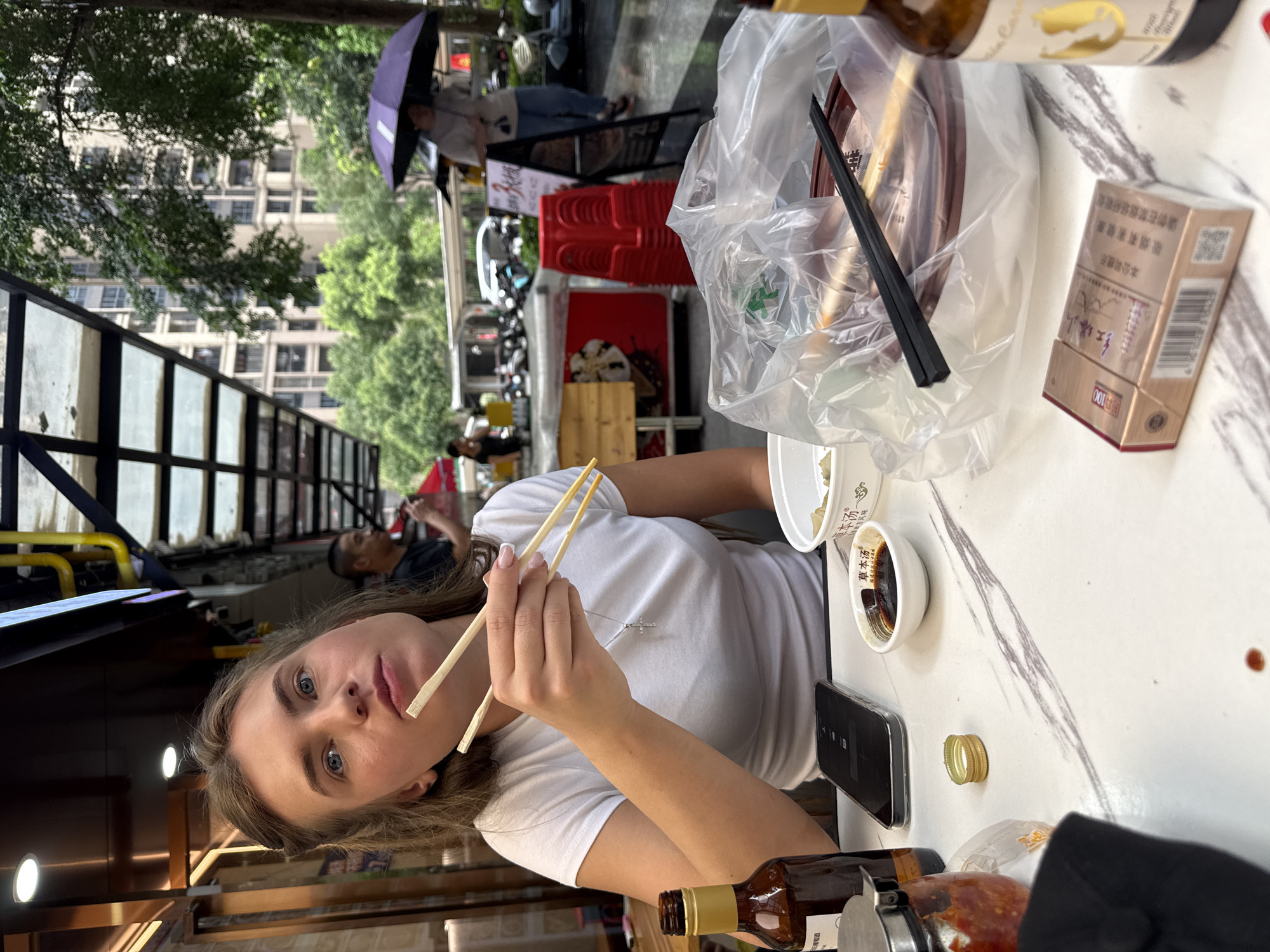 A young Caucasian woman with blonde hair pulled back is sitting at a table outdoors, possibly on a sidewalk cafe in Haizhu District, China. She is wearing a white t-shirt and holding a pair of chopsticks, bringing food to her mouth. The table is cluttered with various items, including a bottle of brown liquid, a smartphone, a small dish of dark sauce, a packet of cigarettes, a bowl of food covered in plastic wrap with black chopsticks resting on top, and another bottle with a gold label. In the background, other people, trees, buildings, and parked motorcycles are visible, suggesting an urban environment. The lighting indicates it is daytime.