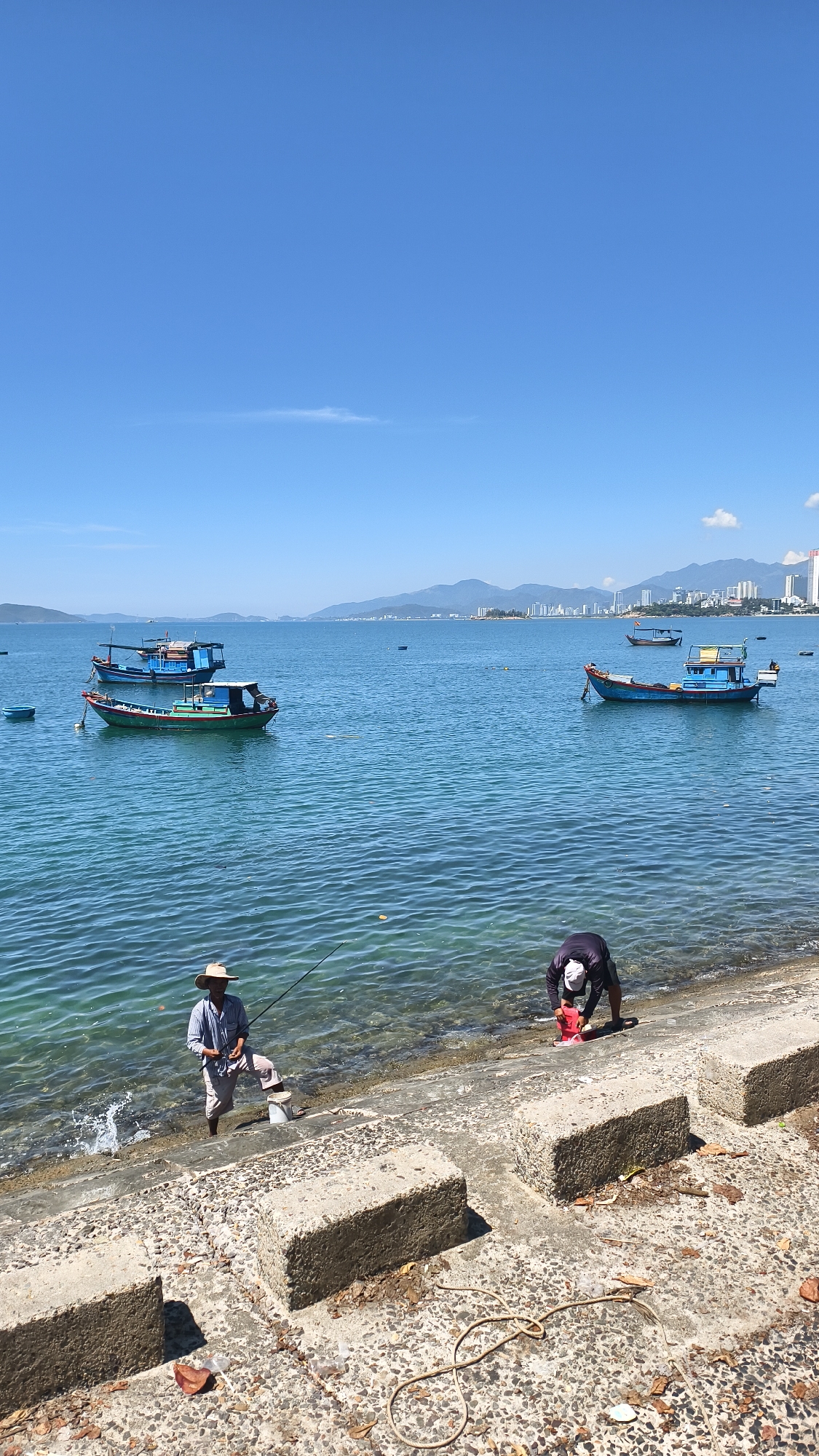 In the city of Bac Nha Trang Ward, Vietnam, a bright, clear day unfolds over a serene seascape. The tranquil azure water laps gently against a concrete embankment, where two individuals are engaged in fishing. To the left, a man in a straw hat and light-colored shirt stands ankle-deep in the water, holding a fishing rod. Beside him, a white bucket rests on the embankment. To the right, another man, dressed in dark clothing, bends over the edge of the embankment, interacting with a bright red object near the water's surface. The embankment itself is constructed from large, rough-hewn concrete blocks. Scattered on the embankment are dried leaves and a coiled rope. In the distance, several fishing boats bob on the water. Beyond the water, a cityscape with numerous buildings and mountainous terrain stretches across the horizon under a vast, cloudless blue sky.