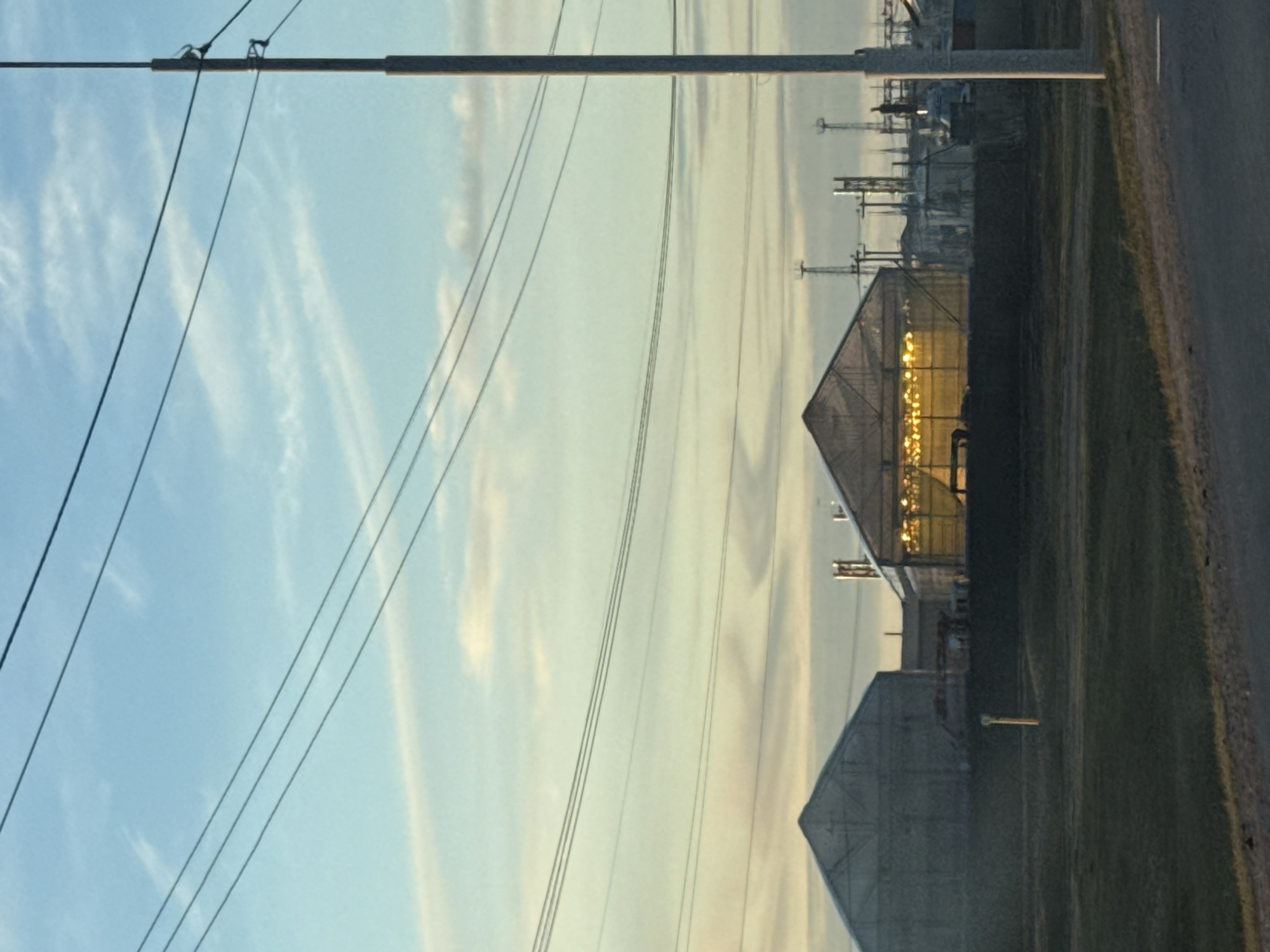 The image shows an industrial landscape in what is purportedly Syrdakh, Russia. A high-voltage power line runs across the top of the frame. Below it, a collection of buildings is visible, including greenhouses. The sun appears to be setting or rising, based on the lighting. A road runs along the right side of the image, with a grassy area between the road and the buildings. There is no sign of people.