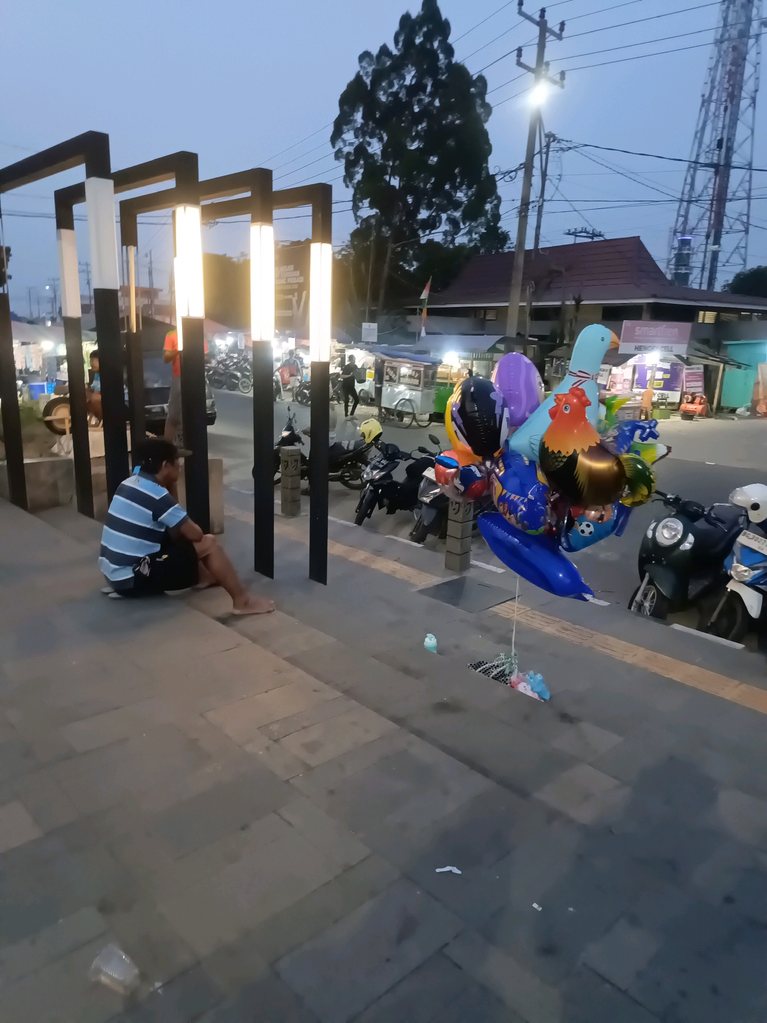 A man sits on the steps of a pedestrian walkway in Lubuklinggau, Indonesia. He is wearing a blue and white striped t-shirt and black shorts, and is barefoot. The walkway is paved with gray tiles. In the background, a collection of colorful balloons, including a blue bird, a red rooster, and several animal-shaped balloons, are tied to a stand. Several motorcycles are parked nearby. The scene is illuminated by streetlights and the fading light of dusk. A tall cell tower is visible in the distance.