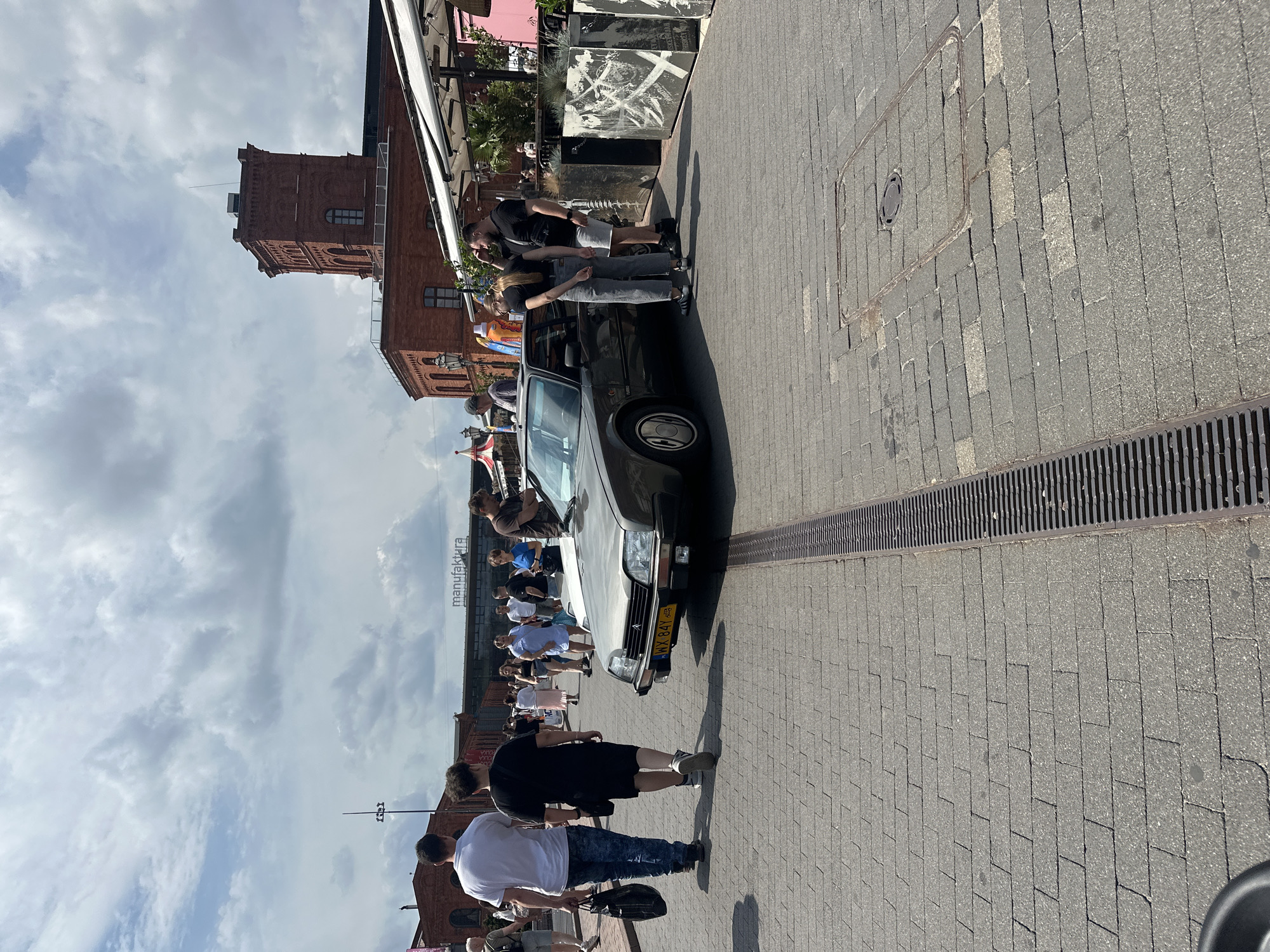 The image captures a sunny day scene in the Manufaktura complex in Łódź, Poland. In the foreground, a vintage gray car with a yellow license plate (WX 84Y) is parked on a cobblestone pedestrian area. Several people are gathered around or walking nearby.
On the left, two men walk away from the camera, one in a white t-shirt and jeans carrying a black bag, and the other in a black t-shirt and shorts. Further back, a group of people are seen strolling, some in casual attire.
The background features the characteristic red brick architecture of Manufaktura, including a prominent red brick tower. White awnings of outdoor seating areas are visible, suggesting a commercial or entertainment district. The sky is partly cloudy. The overall atmosphere is one of a public gathering or event.