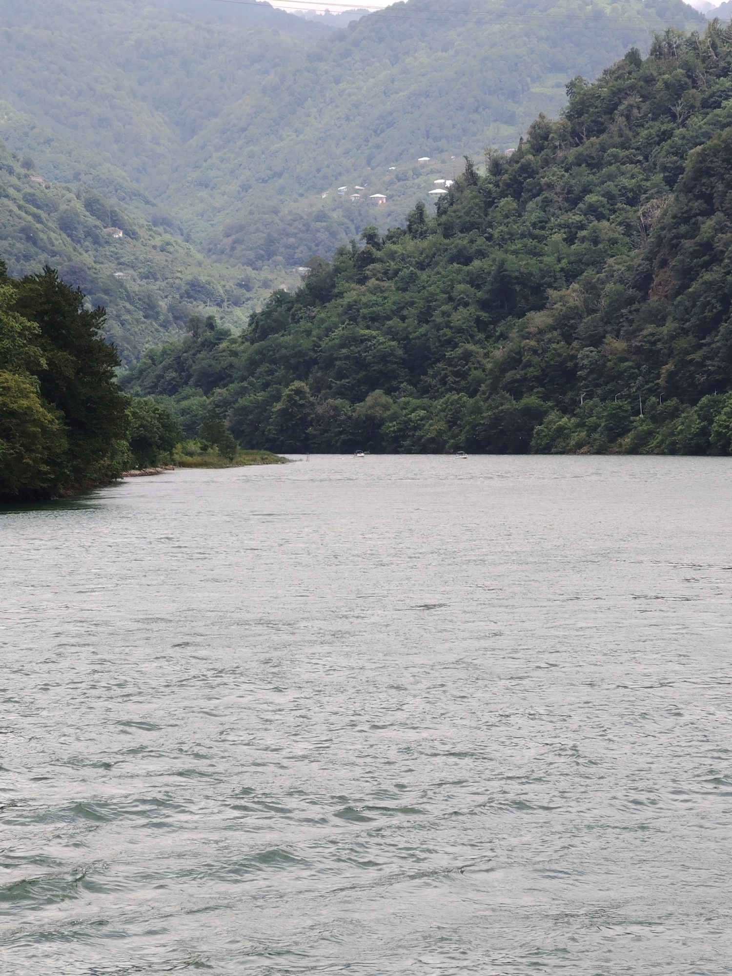 A wide shot shows a calm river in the foreground, reflecting the overcast sky. The river is flanked by dense, green forests that cover steep mountainsides. The mountains rise into the distance, their peaks shrouded in mist. A few small white houses are visible nestled among the trees on the slopes. Two small boats are present on the river. The overall scene is serene and natural, indicative of a rural landscape in Mirveti, Georgia.