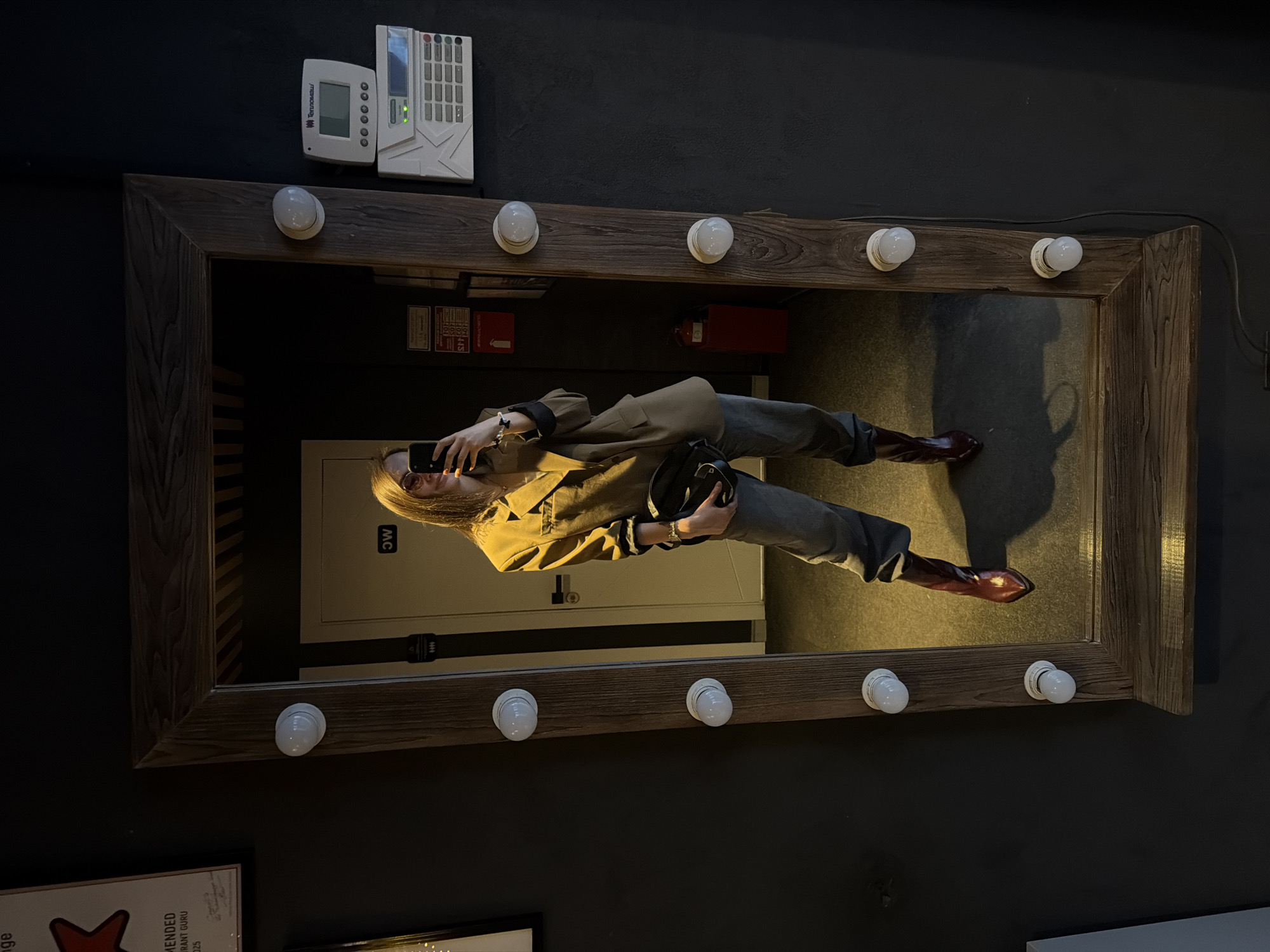 A person is reflected in a mirror with light bulbs around its frame. The person is holding a phone and a black bag, wearing a blazer, jeans, and boots. The mirror hangs on a dark wall, near a digital control panel. The reflection shows a closed door with a sign, and a fire extinguisher. The image was taken in Chaikovsky, Russia.