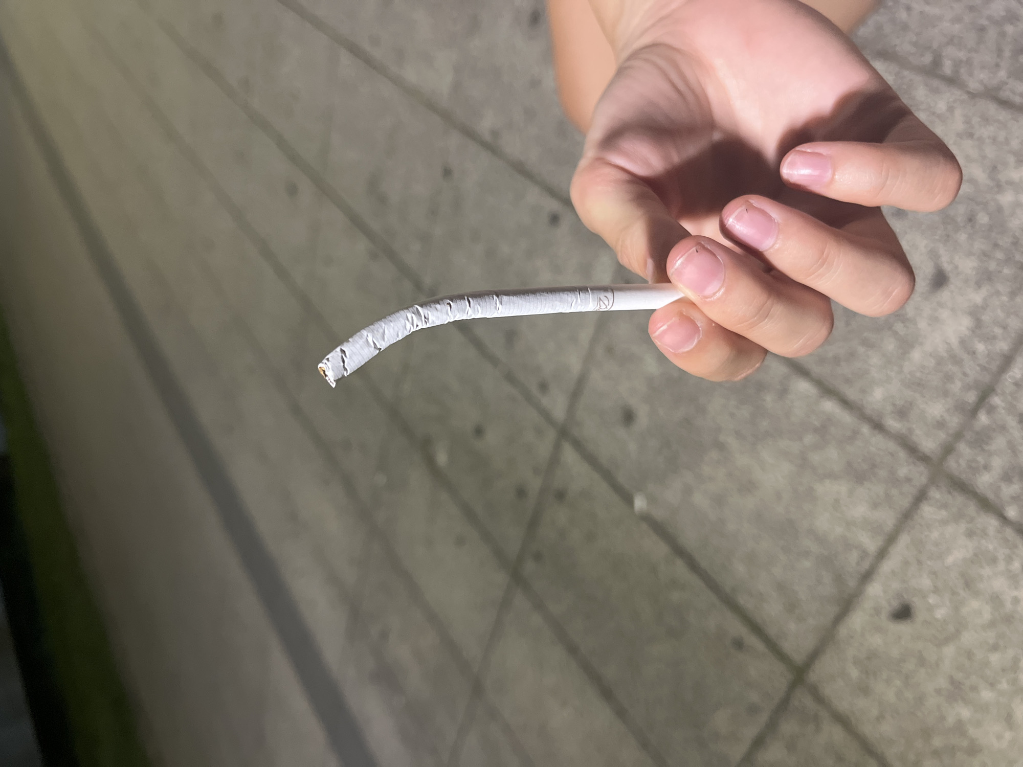 A hand holds a bent cigarette outdoors on a paved surface. The cigarette is white with a filter end, and appears to be partially lit or smoked. The hand is light-skinned and the fingernails are short and unpolished. The background consists of a light-colored, textured pavement with visible grout lines, and some darker areas that could be shadows or wet spots. The scene is lit by artificial light, casting shadows. The location is Vratsa, Bulgaria.