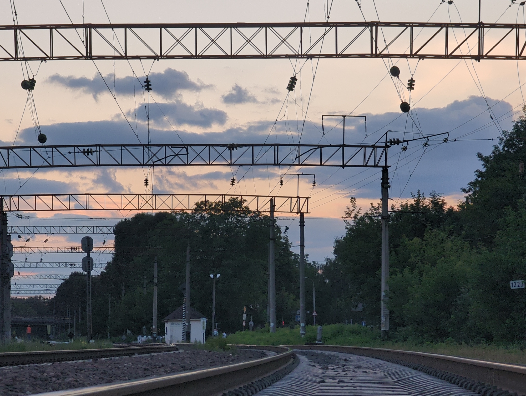 The image depicts a railway track curving to the right, with overhead electrical infrastructure for trains. The sky, tinged with sunset colors, is visible above the metal structures and wires. Dense trees line the right side of the tracks, and a small white building is situated to the left, near a traffic signal. In the distance, more trees and some structures are visible. The overall scene suggests a railway environment at dusk.