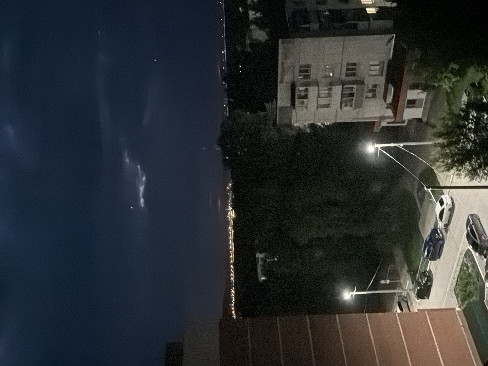 The image depicts a nighttime view of an urban area, likely in Chaikovsky, Russia, characterized by apartment buildings, parked cars, and distant city lights. In the foreground, a portion of a red-brick apartment building is visible on the left. A lit streetlamp stands on a pole near the building, illuminating a parking area with several cars. These include a black sedan, a dark blue SUV, a white hatchback, and a black sedan parked further down. Across the street, a multi-story gray apartment building with numerous windows is visible, some of which are illuminated. Beyond the immediate vicinity of the buildings and trees, a distant cityscape stretches across the horizon, marked by a line of bright lights, suggesting a larger populated area. The sky above is dark blue, with faint clouds and what appear to be two distant celestial bodies or lights. The overall scene conveys a sense of a quiet evening in a residential district.