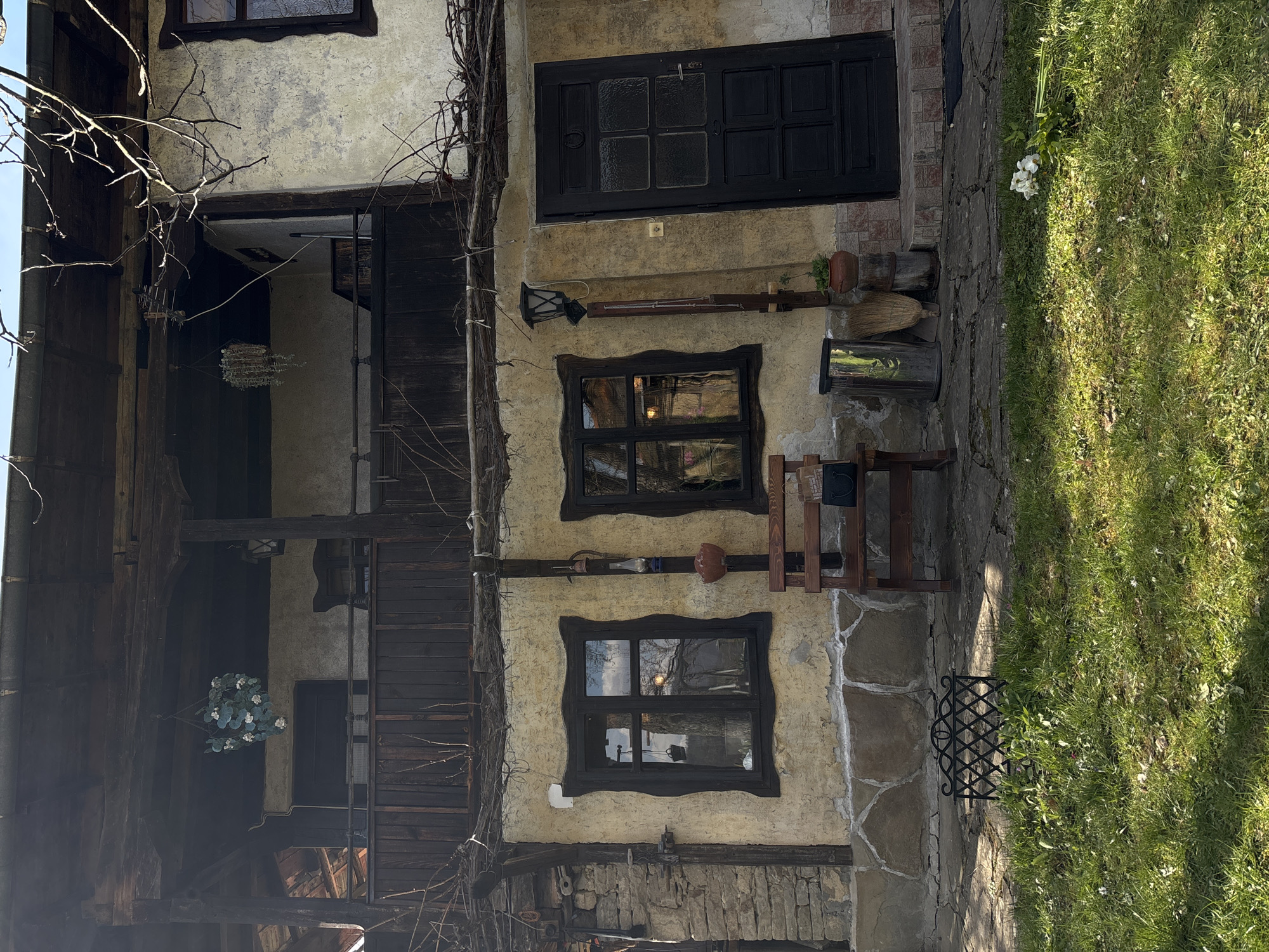 The image captures a building facade in the village of Gorno Trape, Bulgaria. The structure exhibits an old-world charm with its stucco walls, dark wood trim, and aged stone foundation. Two distinct windows with uniquely shaped dark wood frames punctuate the wall, reflecting the outdoor scenery. Above the windows, a dark wood door provides access to the building, while below, a wooden chair and various tools and containers are placed against the wall. To the left of the building, a partial view of a timber-framed structure is visible, adorned with a hanging floral arrangement. On the right, a stone pathway transitions into a grassy area with scattered white flowers, creating a contrast between the man-made structure and the natural environment.