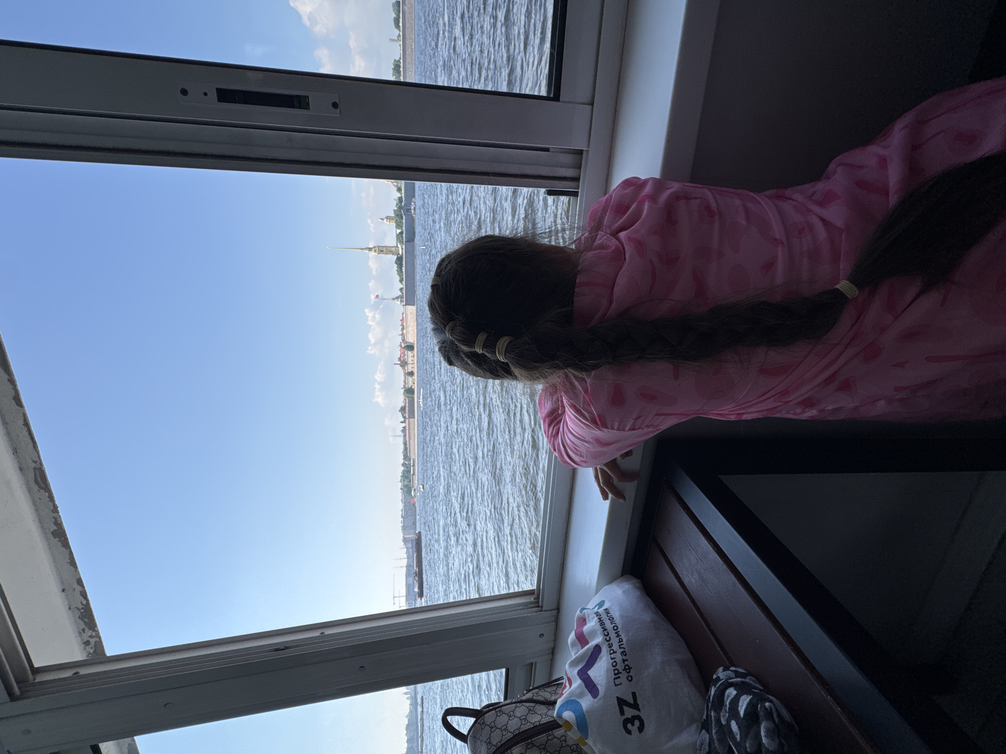 A young girl with dark hair in a braid, wearing a pink patterned garment, looks out a window at the Neva River. In the distance, the Peter and Paul Fortress is visible, with its distinctive spire. The girl is seated on a bench inside a vessel, with a white bag and a backpack on the bench beside her. The sky is blue with scattered clouds.