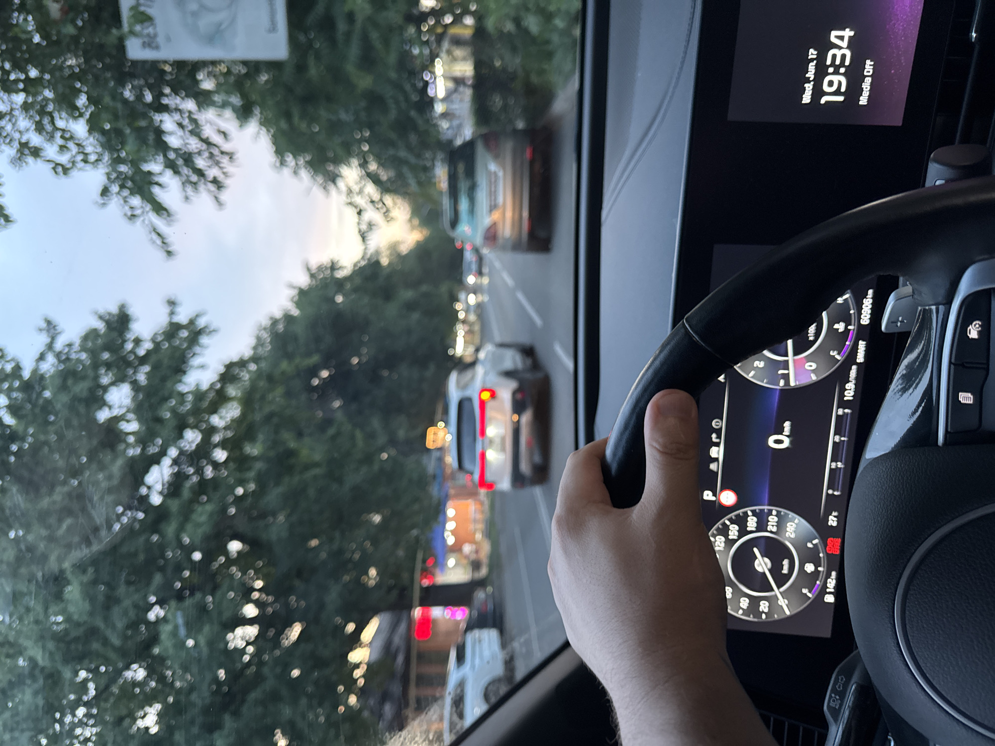 The image is a first-person perspective from inside a car driving on a tree-lined street in Almaty, Kazakhstan. A hand is visible gripping the steering wheel, which has a digital dashboard display. The dashboard shows the car's speed at 0 km/h, indicating it is either stopped or moving very slowly. The digital clock displays "Wed, Jun. 17 19:34." Outside the car, other vehicles are visible on the road, including a white SUV in front and a silver car ahead. Buildings with illuminated signs are present on the right side of the street. The sky is overcast with the soft light of either dawn or dusk. The presence of lush green trees lining the street and the architectural style of the buildings suggest a location within Almaty.