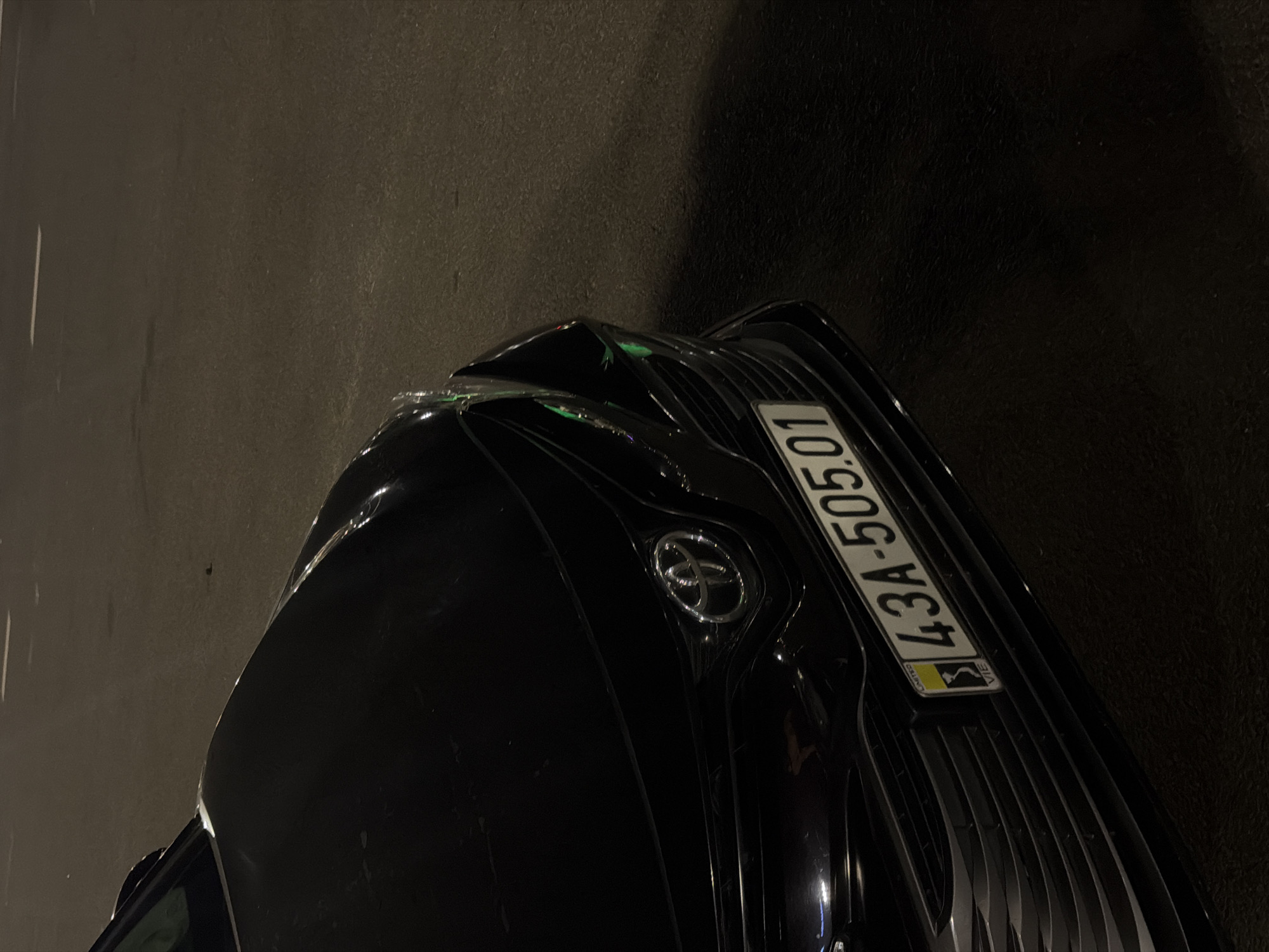The image shows the front of a black car, likely a Toyota Camry, parked on a street at night. The car's license plate, "43A-505.01", indicates it is registered in Đà Nẵng, Vietnam, as "43" is the province code for Đà Nẵng. The license plate also features a smaller yellow and black sticker with "LIMITED" and "VIE" which likely signifies the vehicle type or registration category. The car is parked on asphalt, with faint white lane markings visible in the background. The lighting suggests artificial streetlights or the car's headlights illuminating the scene. The overall environment is dark, typical of a street at night in an urban area. There are no people or other vehicles clearly visible in the frame.